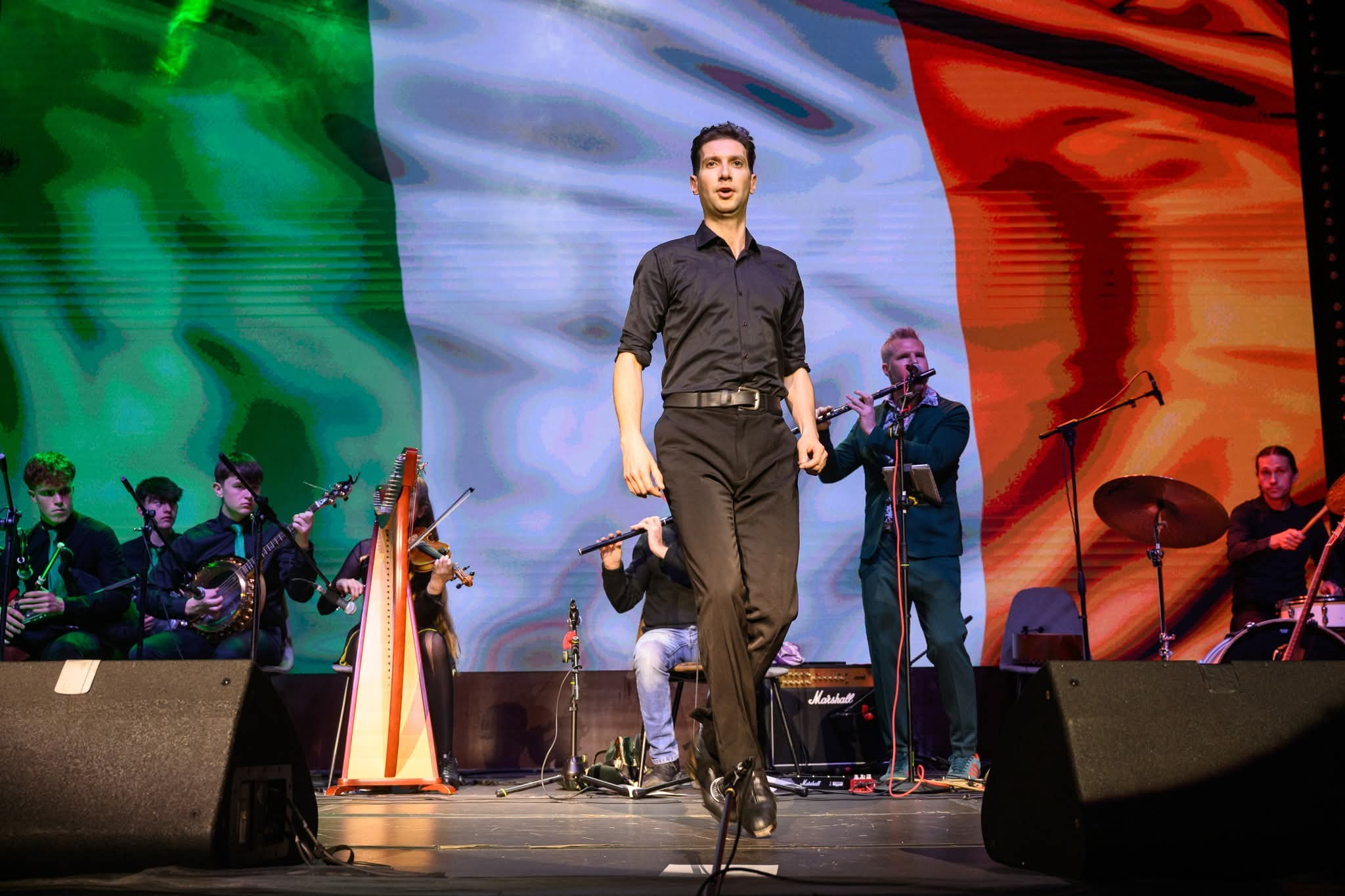 A man in a black shirt and black pants standing on stage singing or speaking, with a large Italian flag as the backdrop. Several musicians are seated on stage playing instruments, and a person is playing a flute behind the man. Audio equipment and a Marshall amplifier are visible in the foreground.