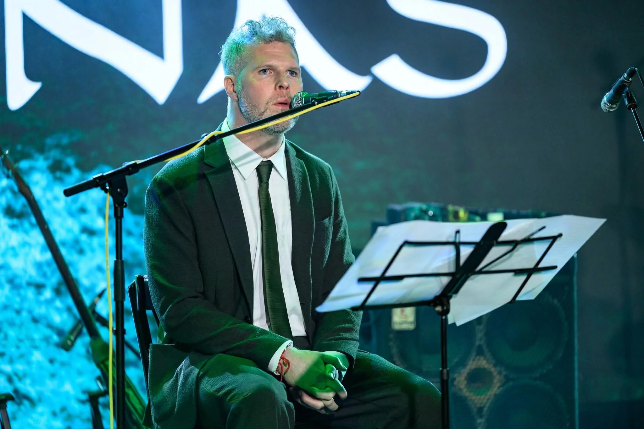 A man in a suit and tie sitting in front of a music stand with sheet music, performing or rehearsing on stage with microphones and stage lighting.