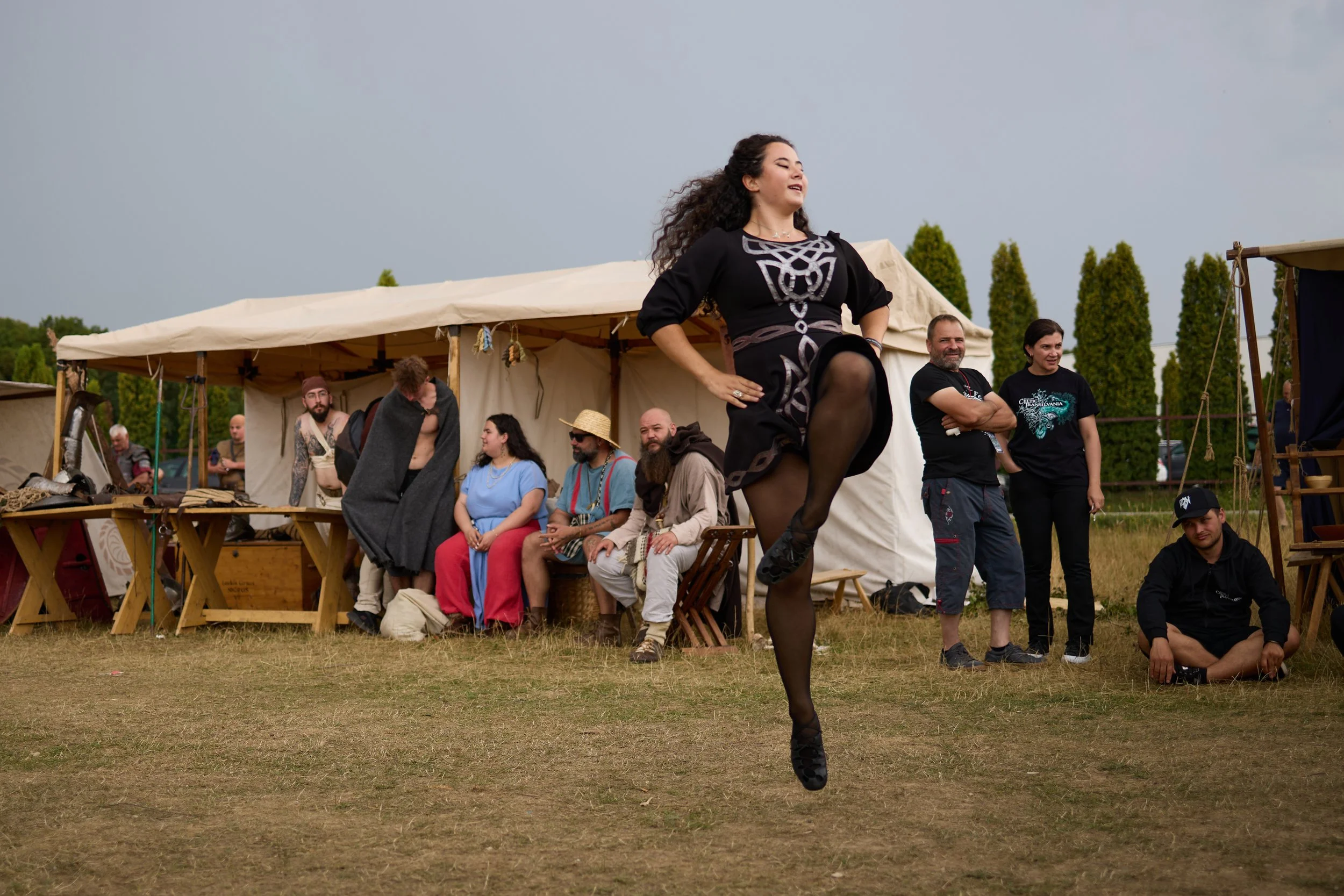 A woman in black with intricate silver and white patterns on her dress is dancing or performing in front of an audience outdoors. The audience, sitting and standing, is casually dressed and appears to be watching her performance. In the background, t