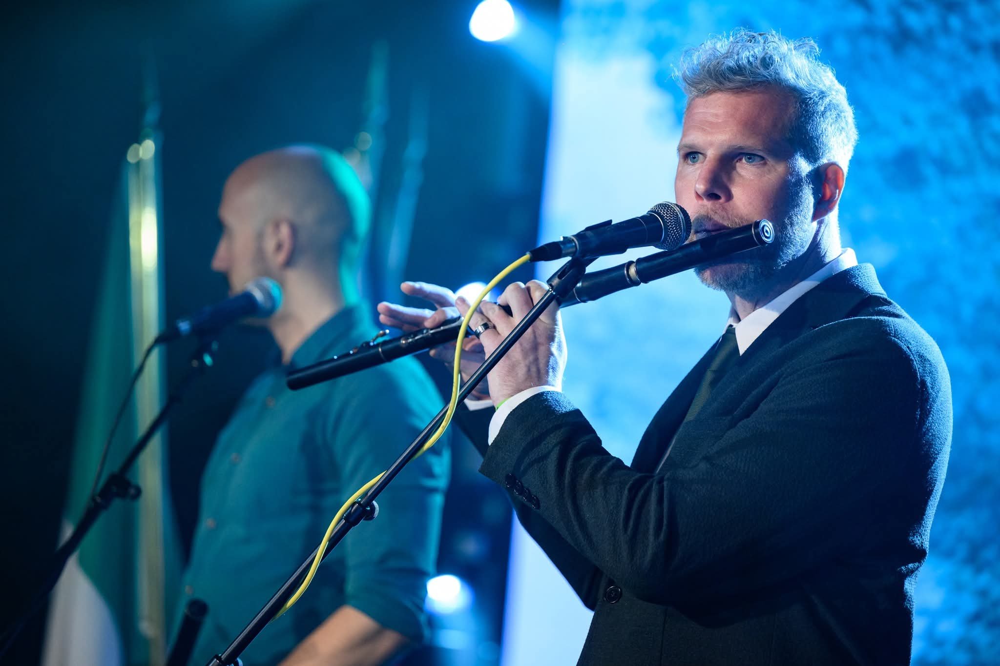 A man with gray hair playing a flute on stage during a performance, with another musician blurred in the background, under blue stage lighting.
