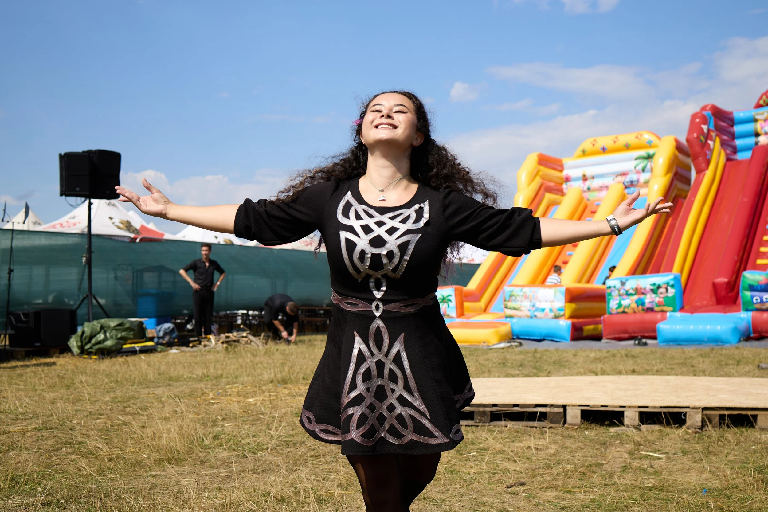 A woman with long curly hair smiling with her arms open wide at an outdoor event, with a colorful inflatable slide and other festival structures in the background under a partly cloudy sky.