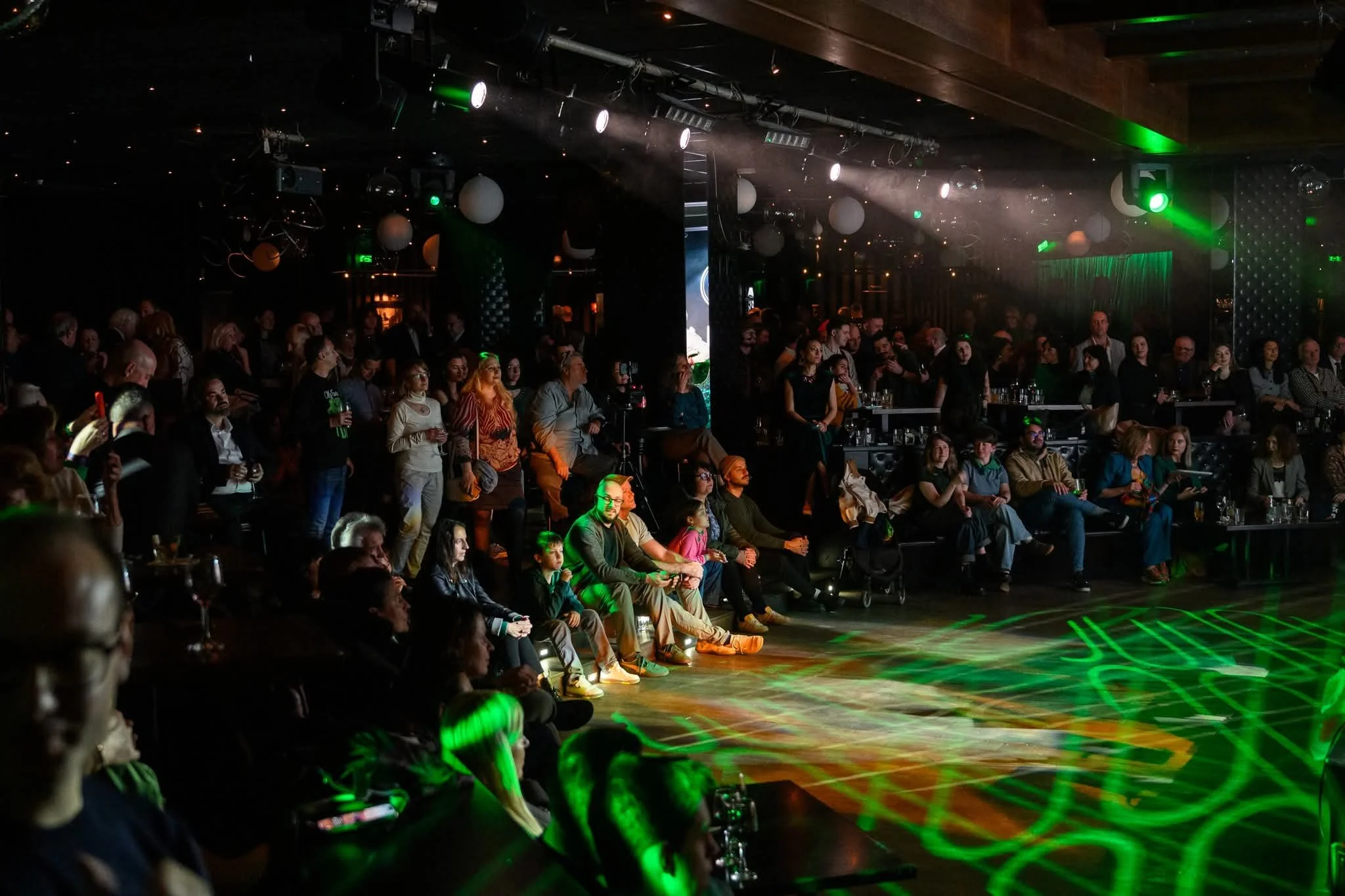 Audience gathered in a dimly lit venue with a stage, illuminated by green and white lights, watching a performance.