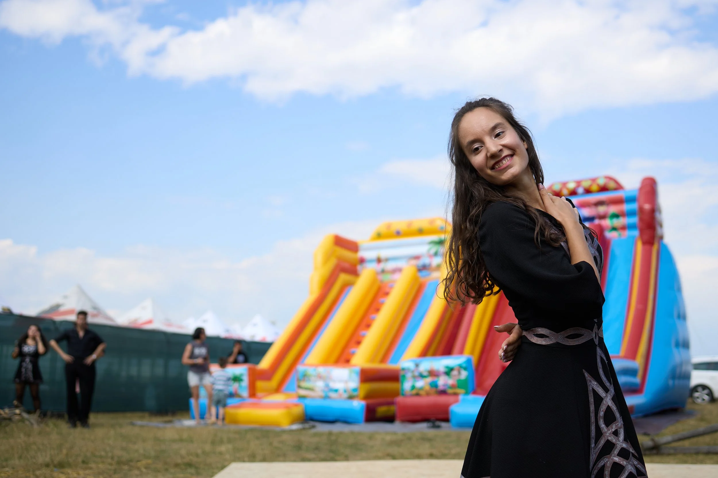A young woman smiling and posing at a fairground, with colorful inflatables and arcade games in the background, under a partly cloudy sky.
