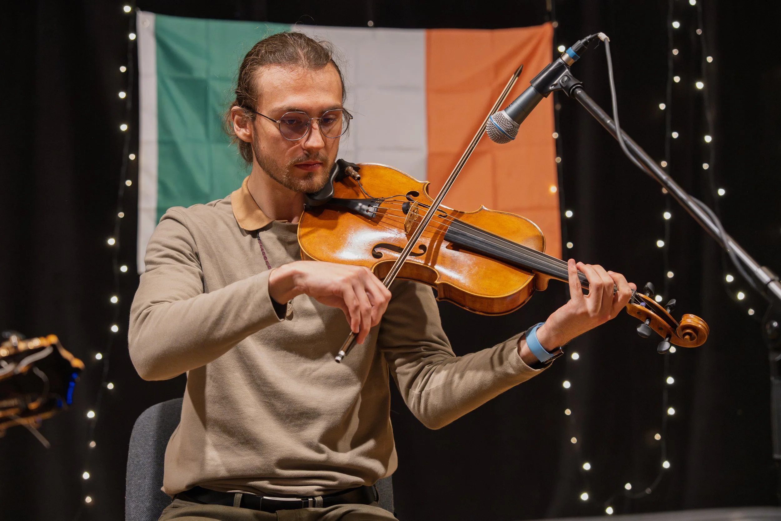 A man playing the violin in front of the Irish flag, with a microphone set up nearby, in a space decorated with string lights.