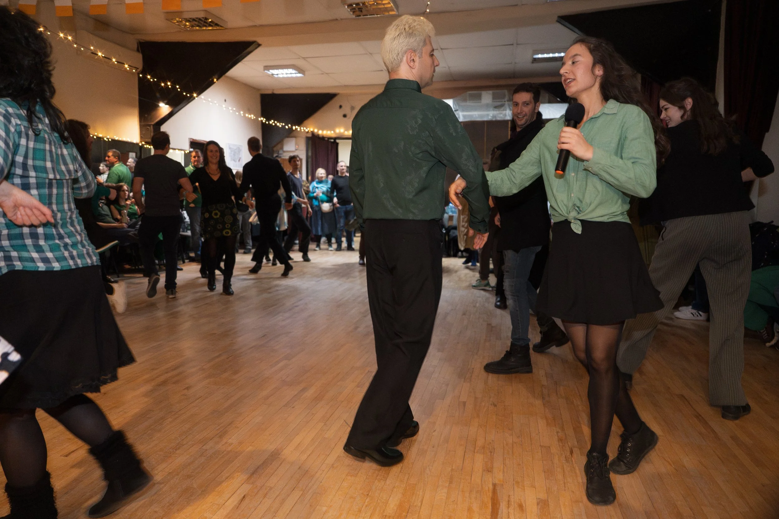 People dancing in a ballroom, with a woman holding a microphone and a man in the foreground. Others in the background are socializing and enjoying the event, with string lights hanging from the ceiling.