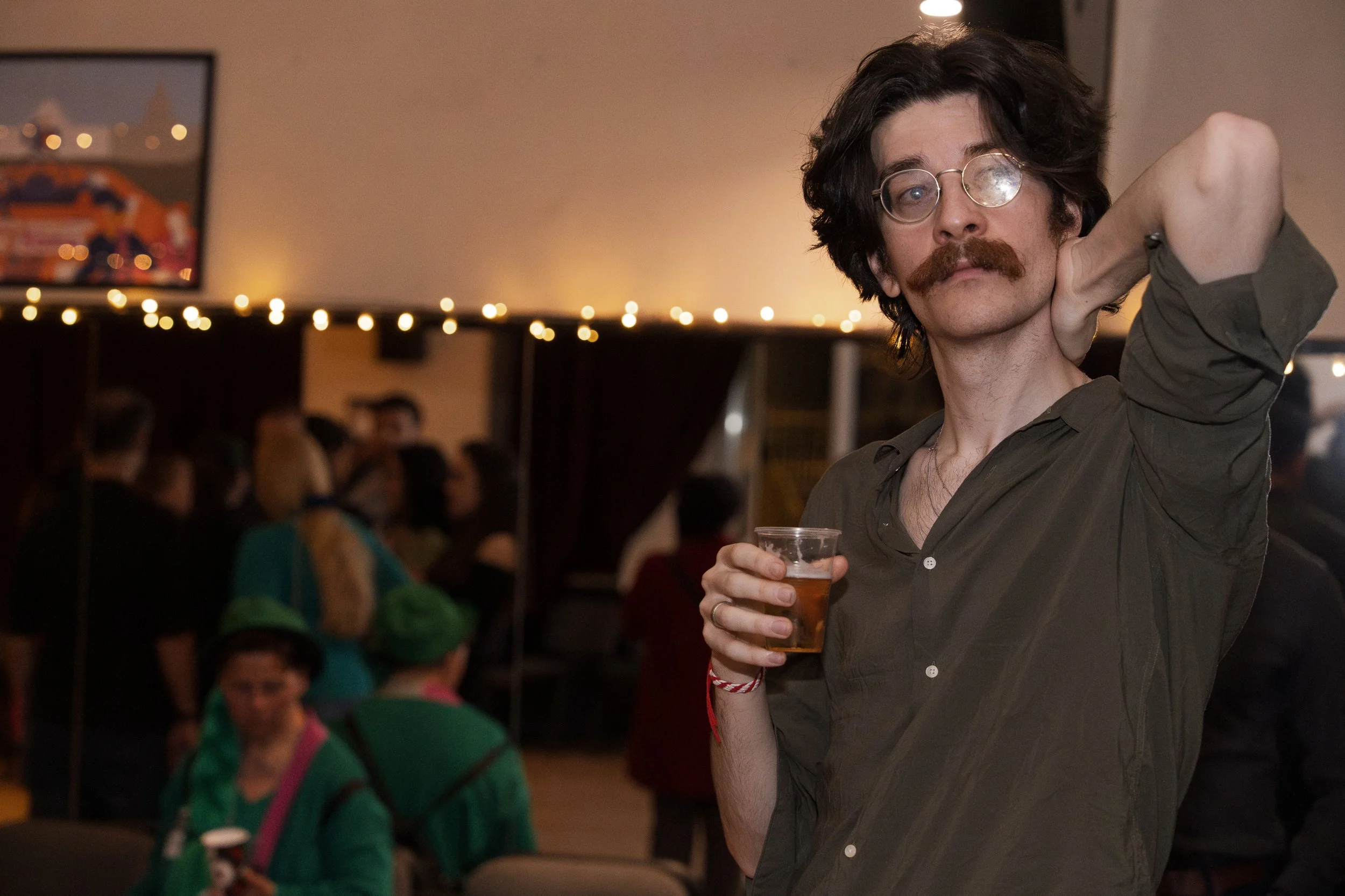 A man with dark, wavy hair, glasses, and a mustache holding a drink in a clear plastic cup, raising his arm with his elbow bent in a social setting with blurred people and warm lighting.