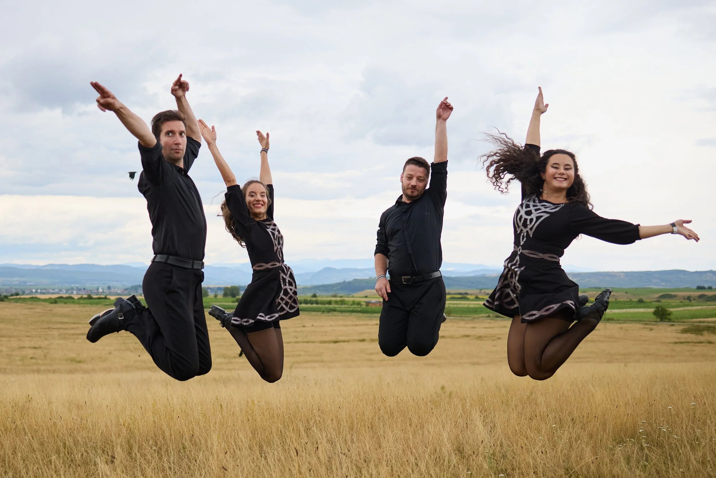 Four people in black outfits jumping joyfully in a field with a scenic landscape and cloudy sky in the background.