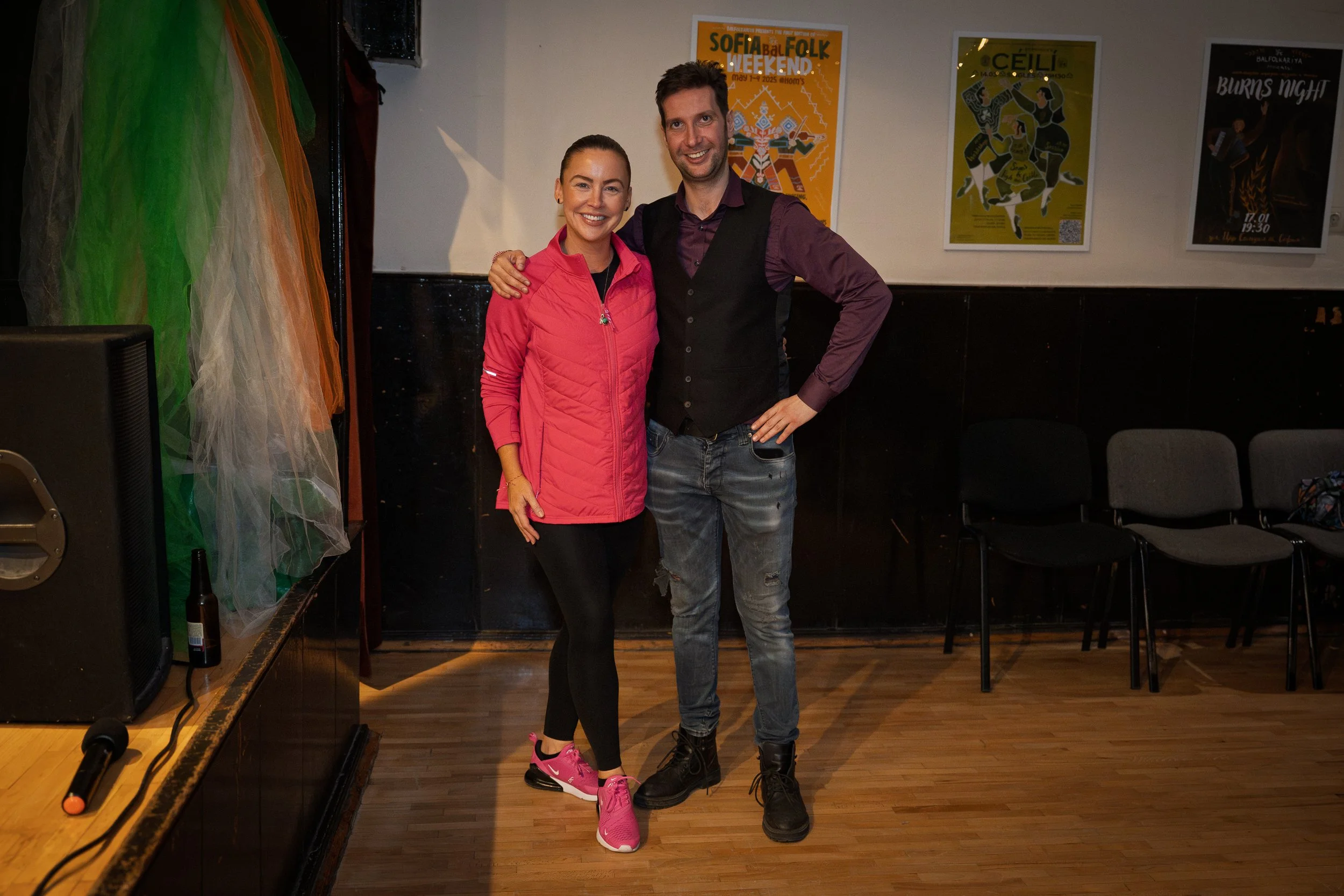 A woman and a man standing closely together, smiling at the camera in an indoor setting. The woman is wearing a pink jacket and pink sneakers, and the man is wearing a dark vest over a maroon shirt with ripped jeans. Behind them are posters on the wa