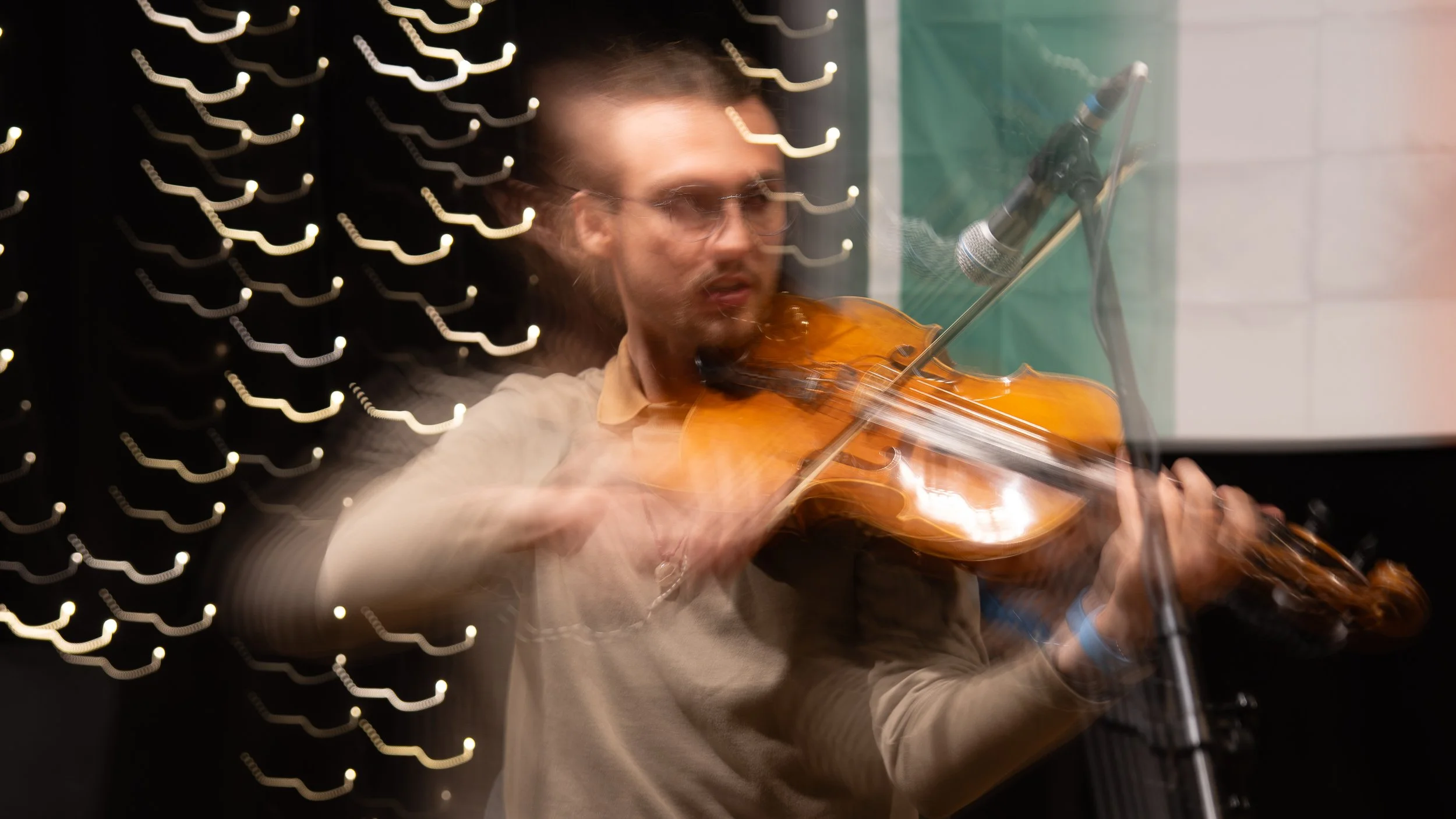 A man wearing glasses and a beige shirt playing a violin with a microphone in front of him, blurred with motion effect, in front of a decorative background with string lights.