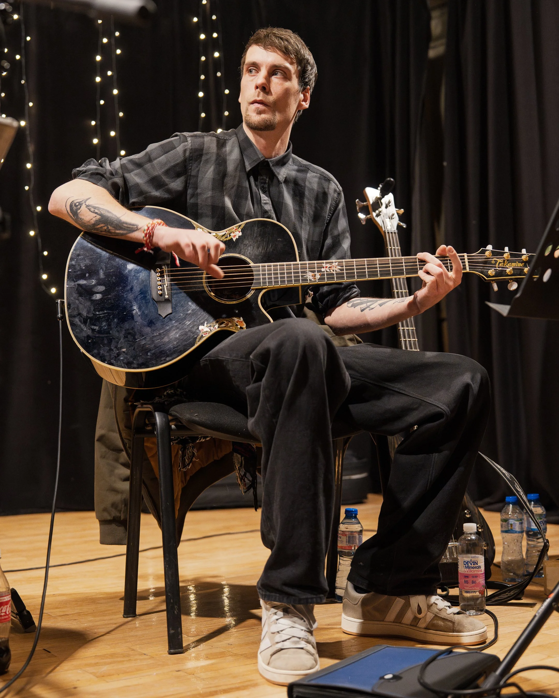 A man with dark hair and tattoos playing an acoustic guitar during a performance or rehearsal on a stage with black curtains and string lights, with water bottles and equipment on the floor.