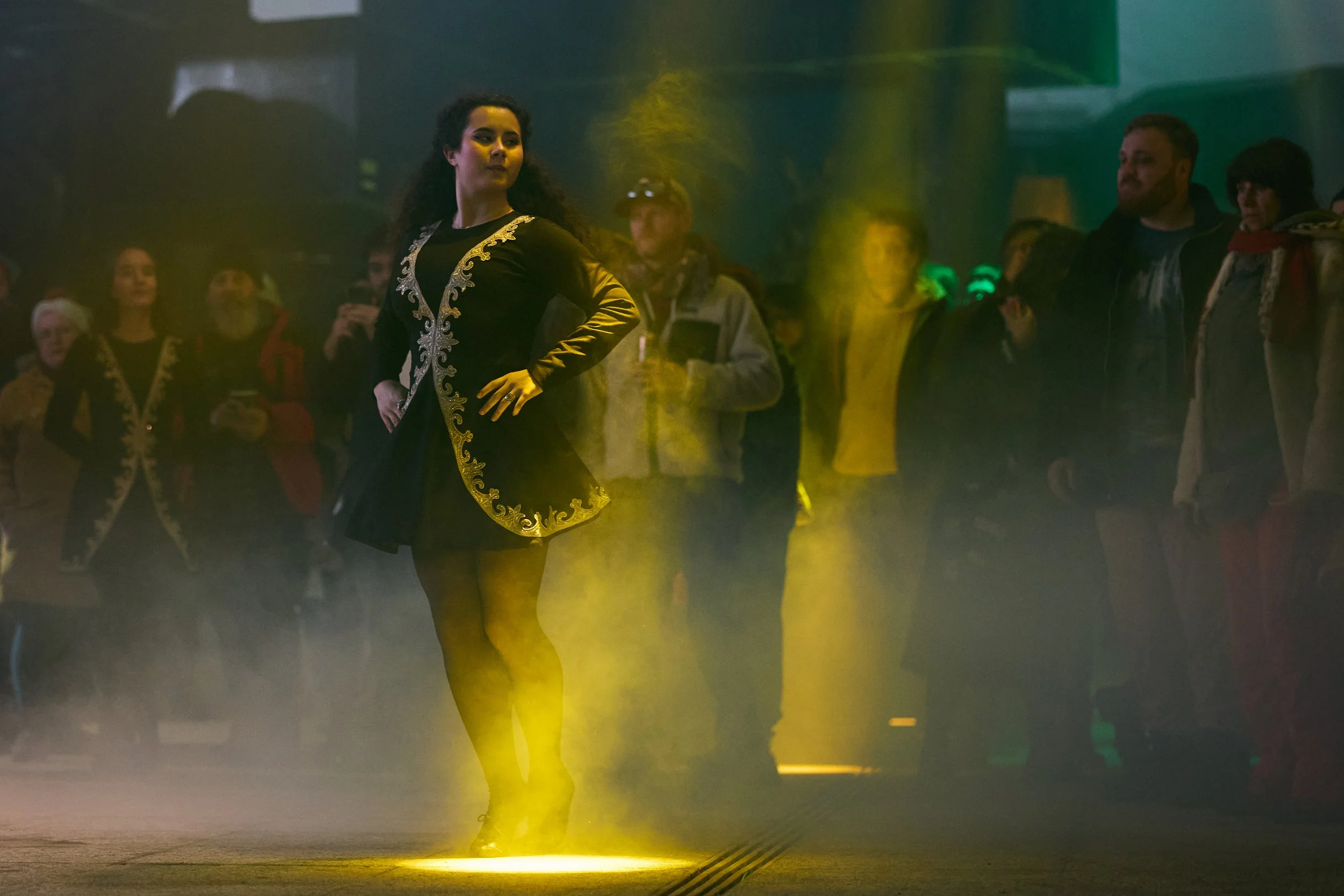 A woman with curly dark hair wearing a black dress with gold embroidery poses with her hand on her hip in a dark, smoky environment with yellow and green lighting, surrounded by onlookers.