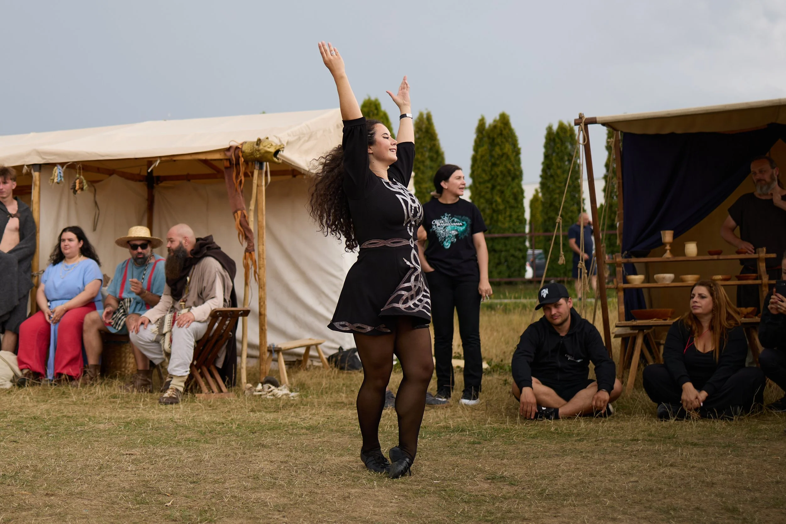 A woman in black dress and tights dancing with her arms raised at an outdoor event, with people seated and standing nearby under tents with green trees in the background.