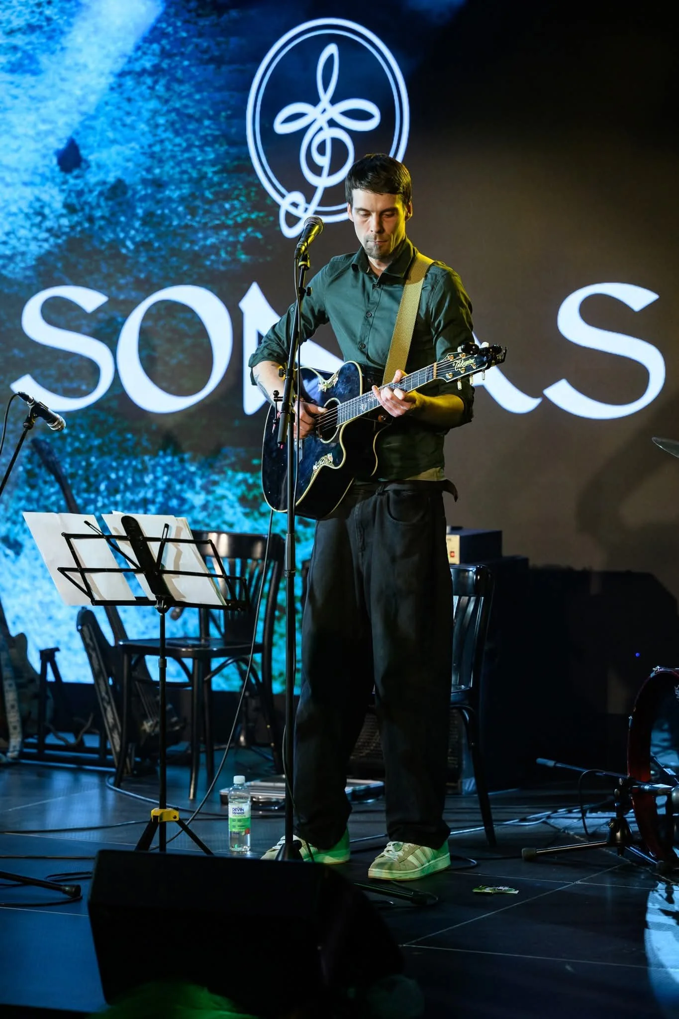A man playing an acoustic guitar on stage with a microphone, music stands, and chairs around him, in front of a screen displaying a logo with a treble clef and stylized loops.