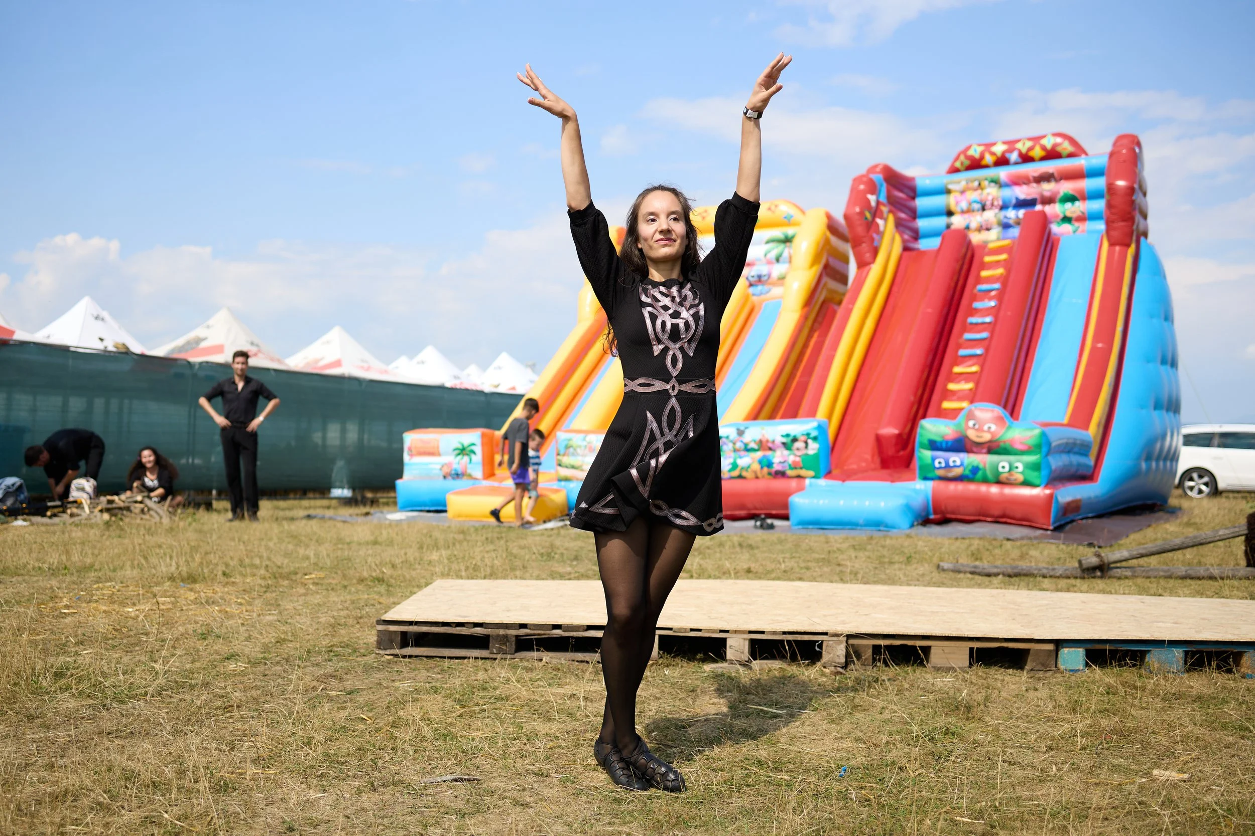 A woman with long hair and black dress standing on a wooden platform at an outdoor amusement park or fair, with colorful inflatable slide and tents in the background.
