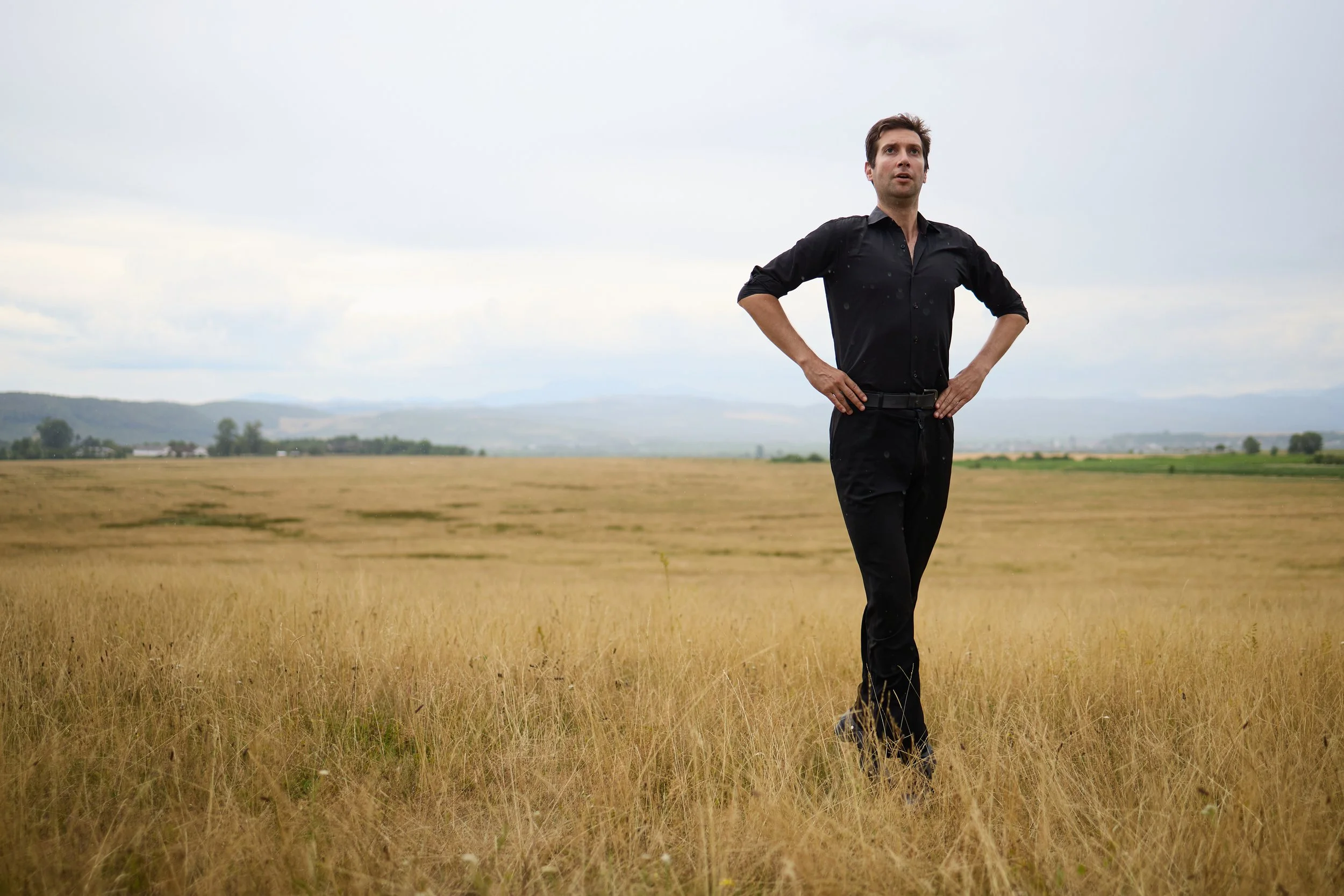 A man in a black shirt and black pants standing in a golden field with his hands on his hips, looking into the distance under a cloudy sky.