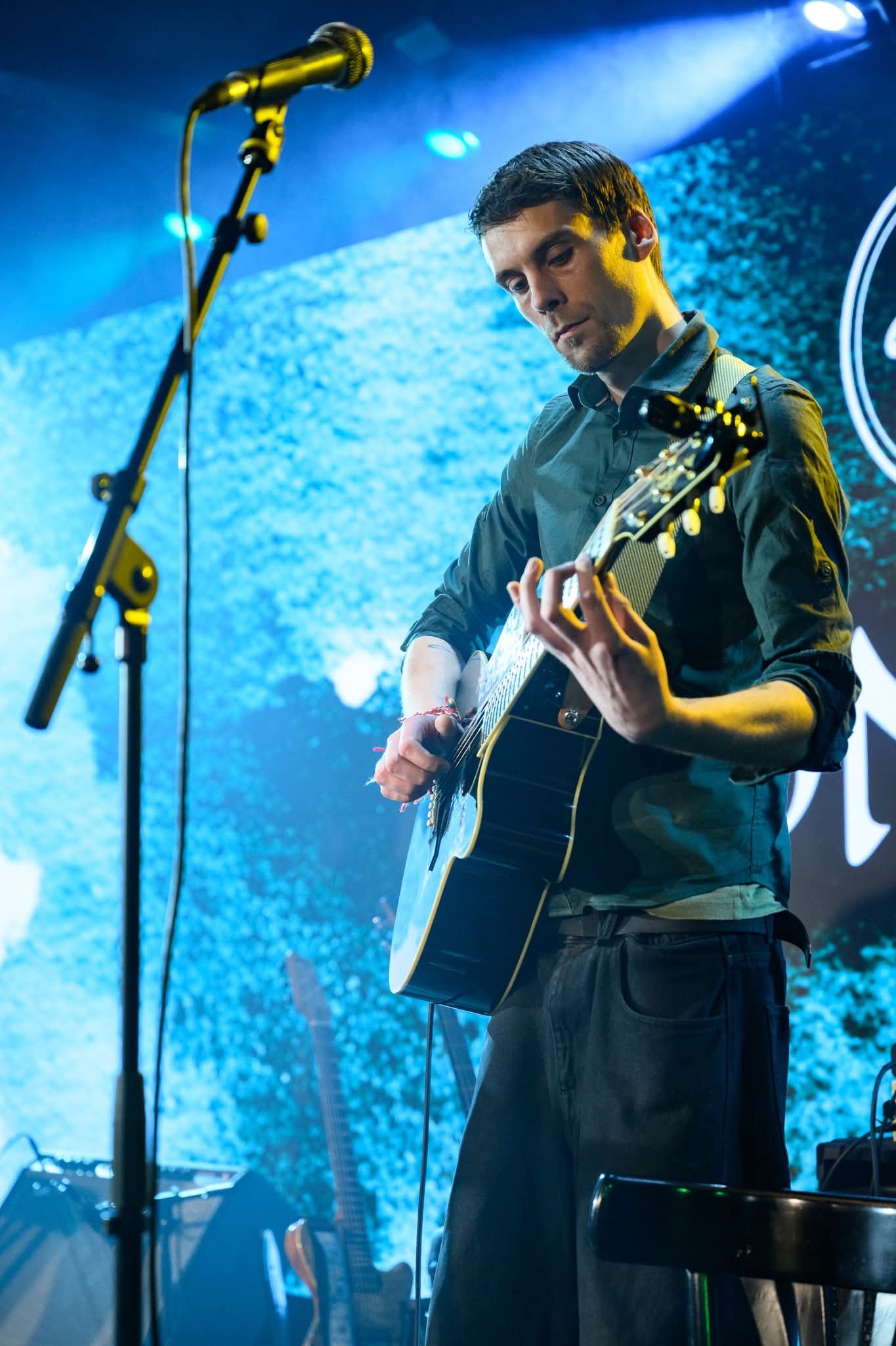 A man playing an acoustic guitar on stage with a microphone in front of him, illuminated by blue and yellow stage lights, in a concert setting.