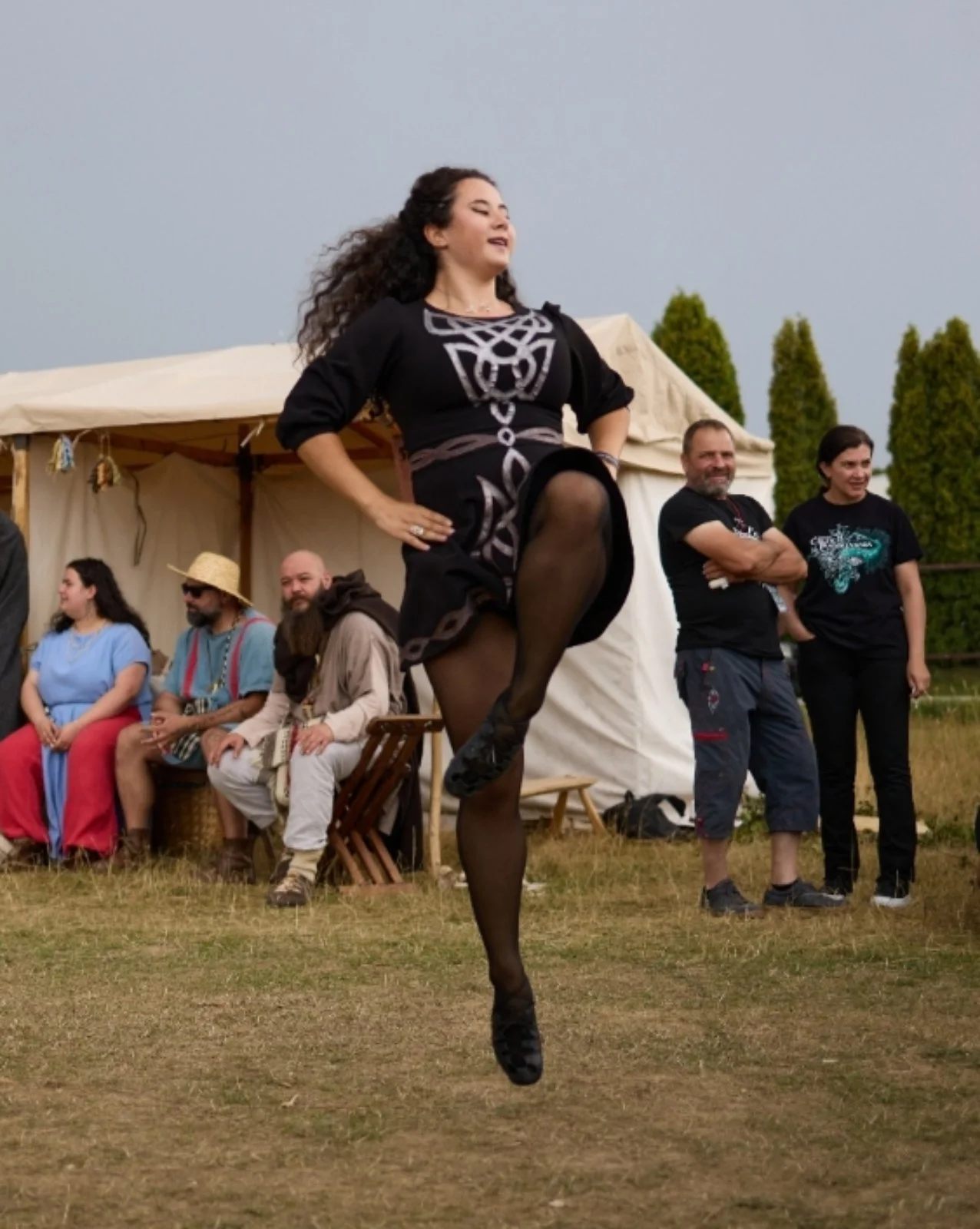 A woman dressed in black with intricate silver patterns is performing a high kick during an outdoor event. Several people are sitting and standing in the background near a white tent, with some watching her performance.