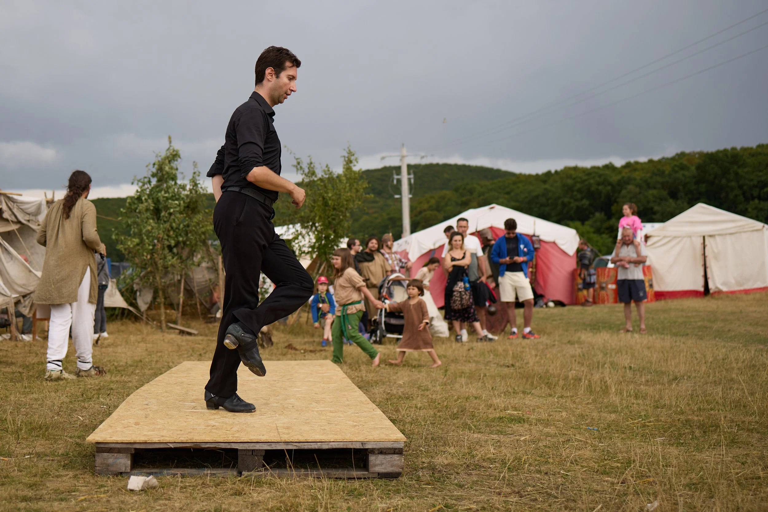 A man dressed in black jumpsuit and boots performs a dance or leap on a small wooden stage outdoors during a gathering with tents and a crowd of people, under a cloudy sky.