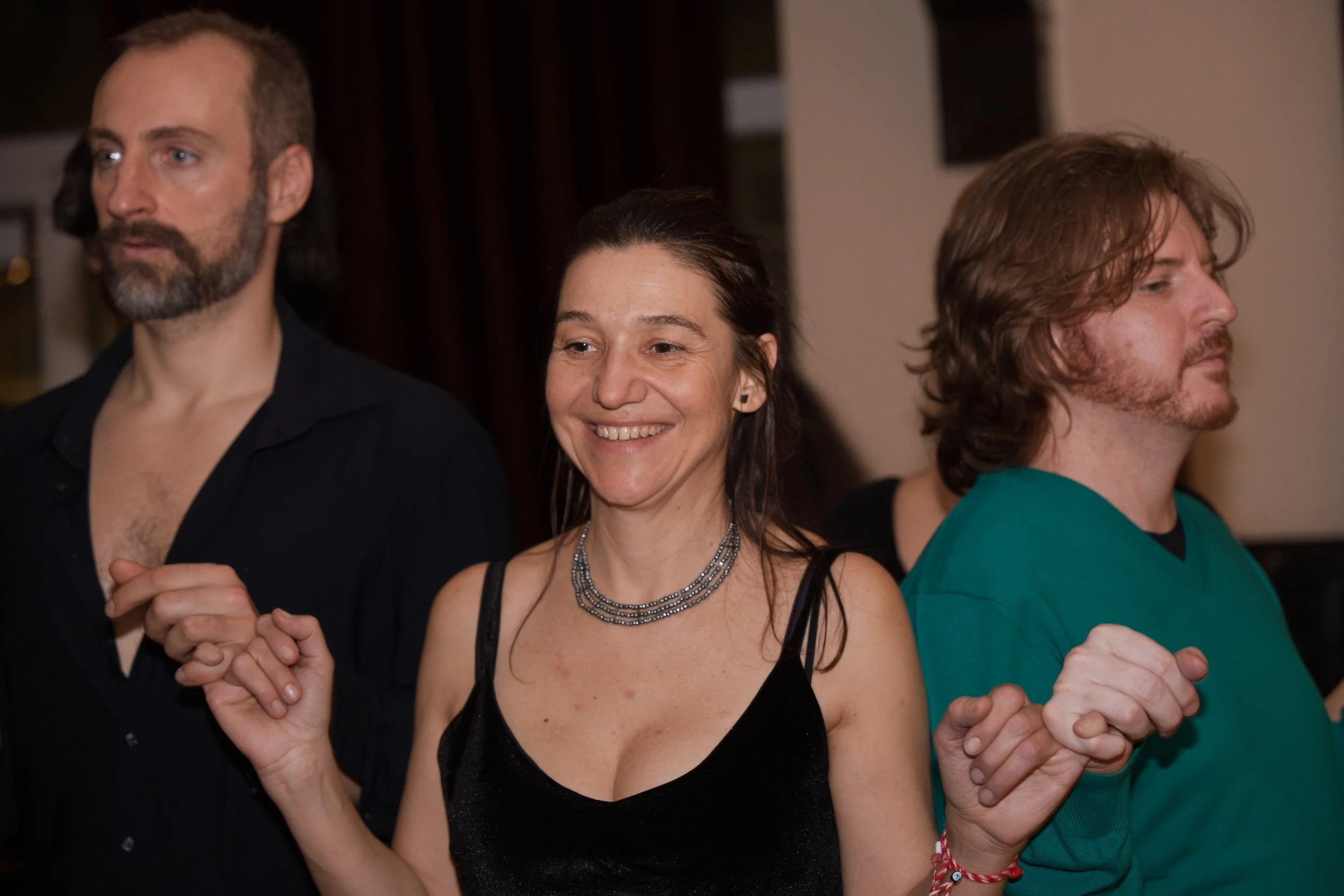 Three people holding hands, standing side by side in a line. The woman in the center is smiling and wearing a black dress and a multi-layered necklace. The man on the left has a beard and is dressed in black, looking to the left. The man on the right
