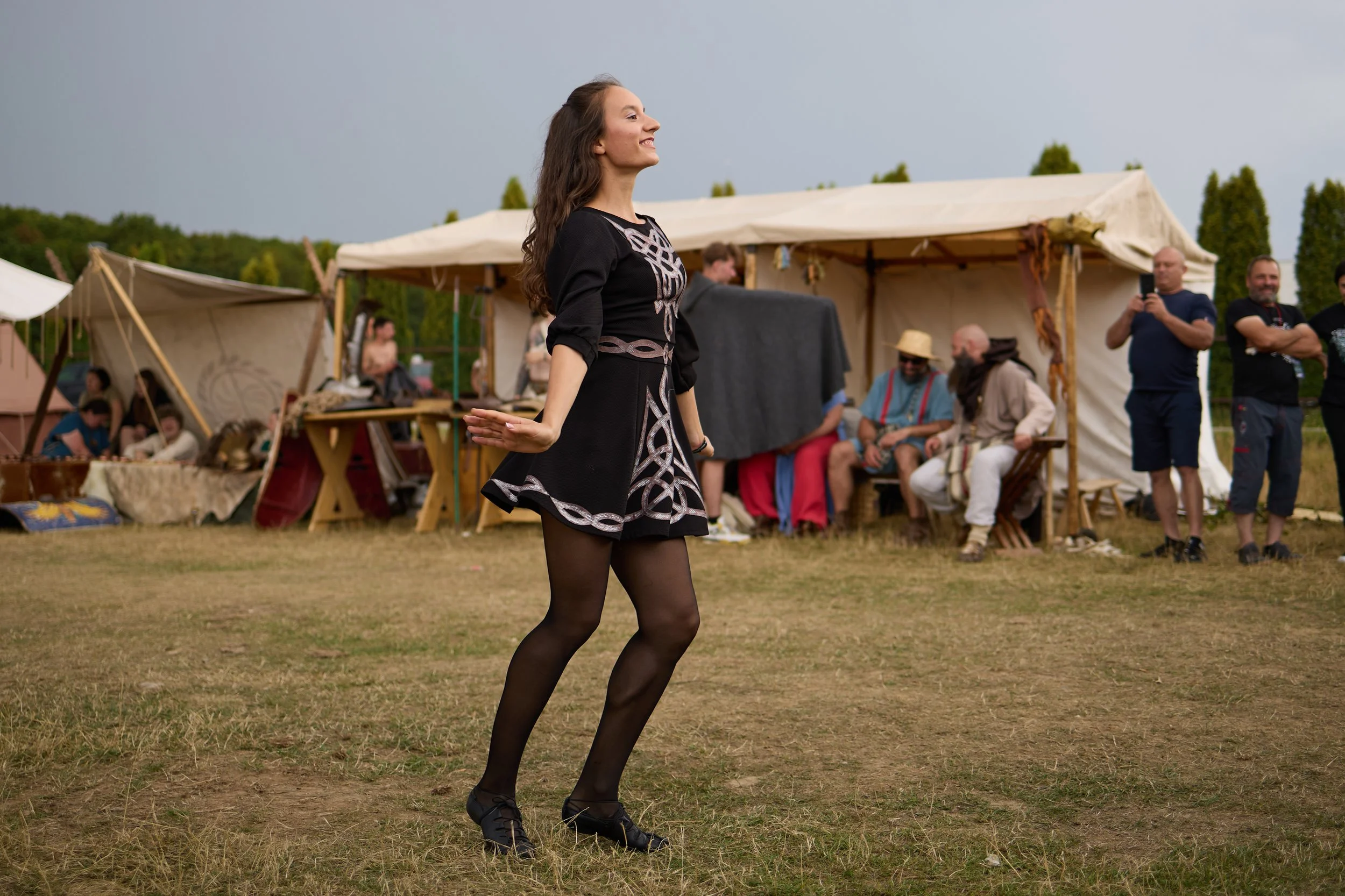 A woman dancing outdoors at a historical reenactment or medieval fair, with tents and spectators in the background.