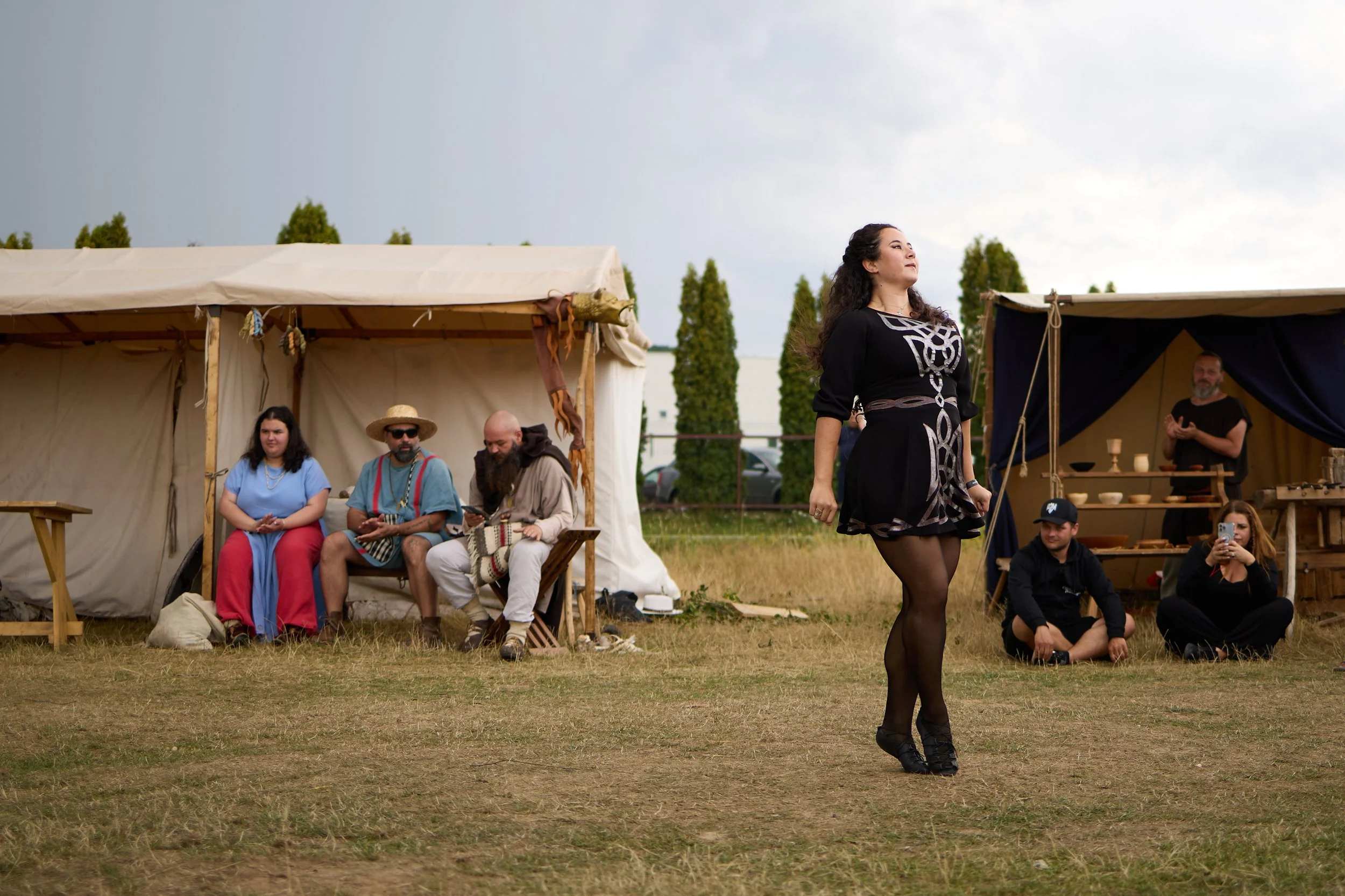 A woman in black dress and tights dancing or performing in front of seated and standing people outdoors during a cloudy day, with tents and trees in the background.