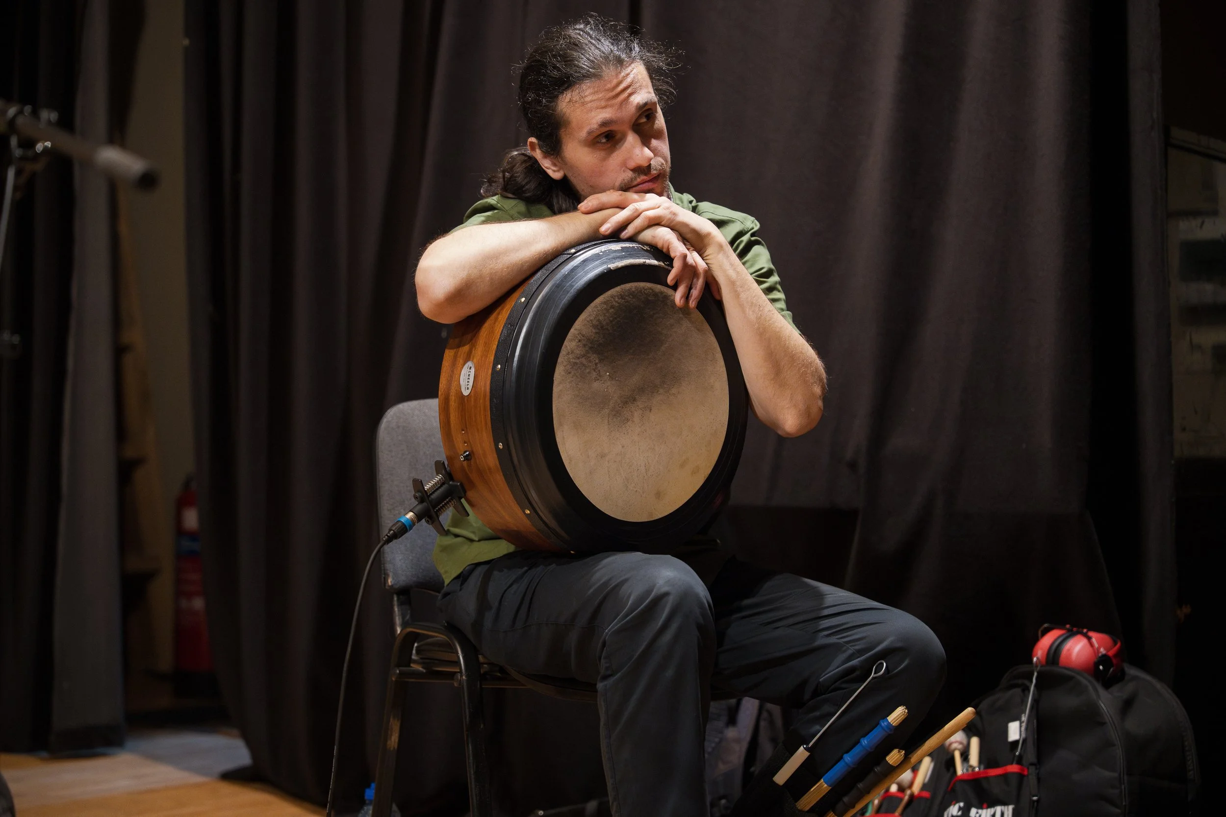 A man with long hair, wearing a green shirt, sitting on a chair, looking thoughtfully while resting his chin on his crossed arms on top of a large, round wooden frame instrument, possibly a drum.