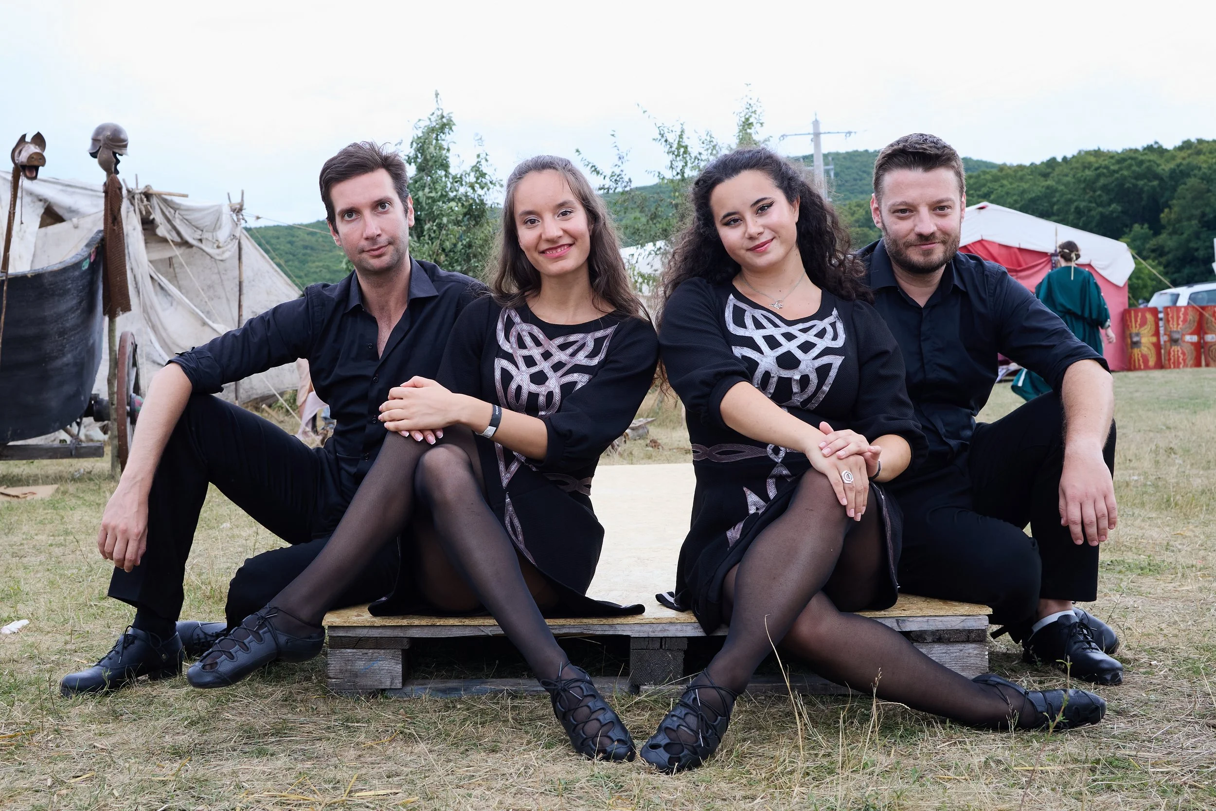 Four people sitting on a wooden platform outdoors, dressed in black, with tents and green hills in the background.