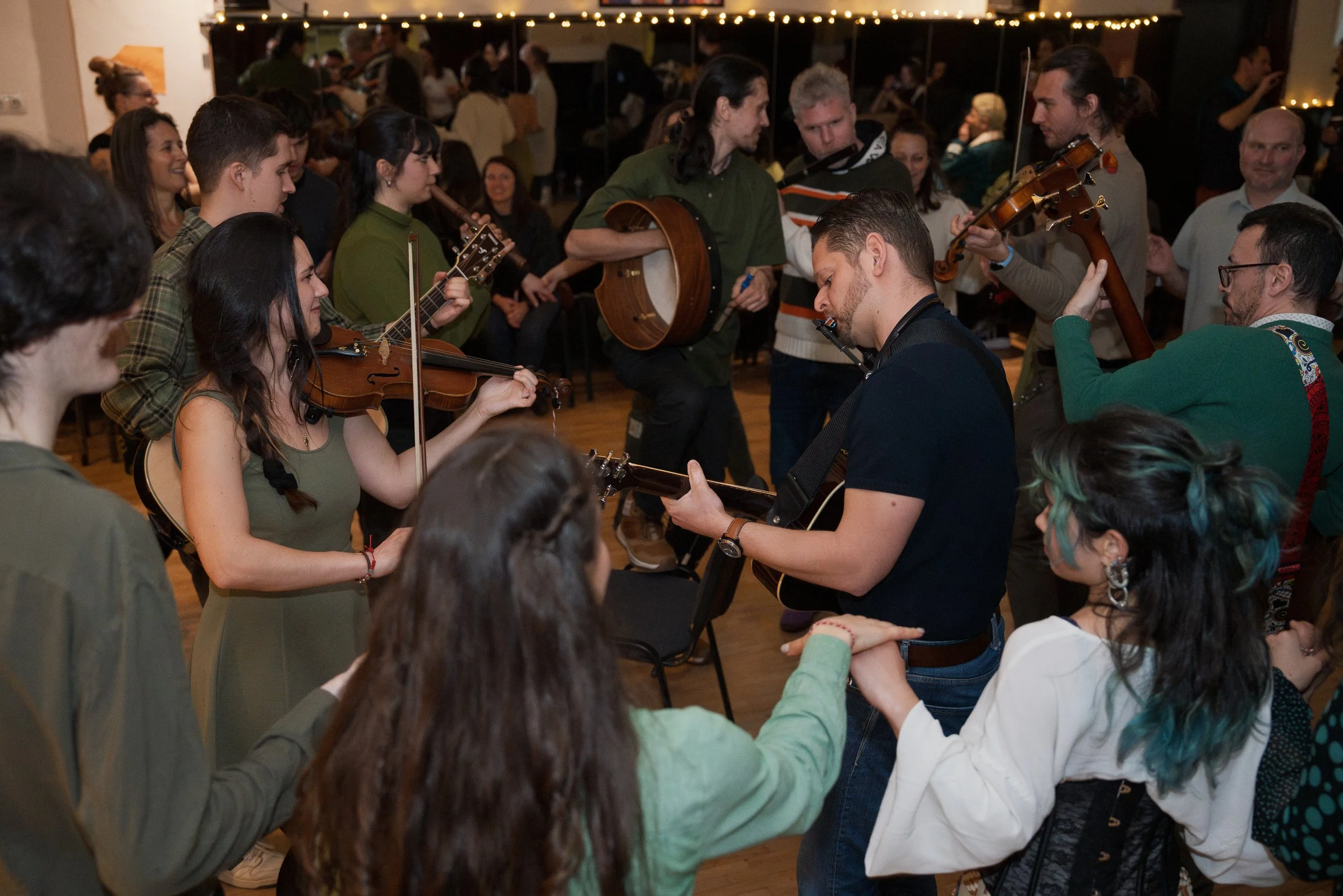 A diverse group of people gathered indoors, some playing musical instruments like guitars and violins, surrounded by onlookers in a cozy, festive setting with string lights overhead.