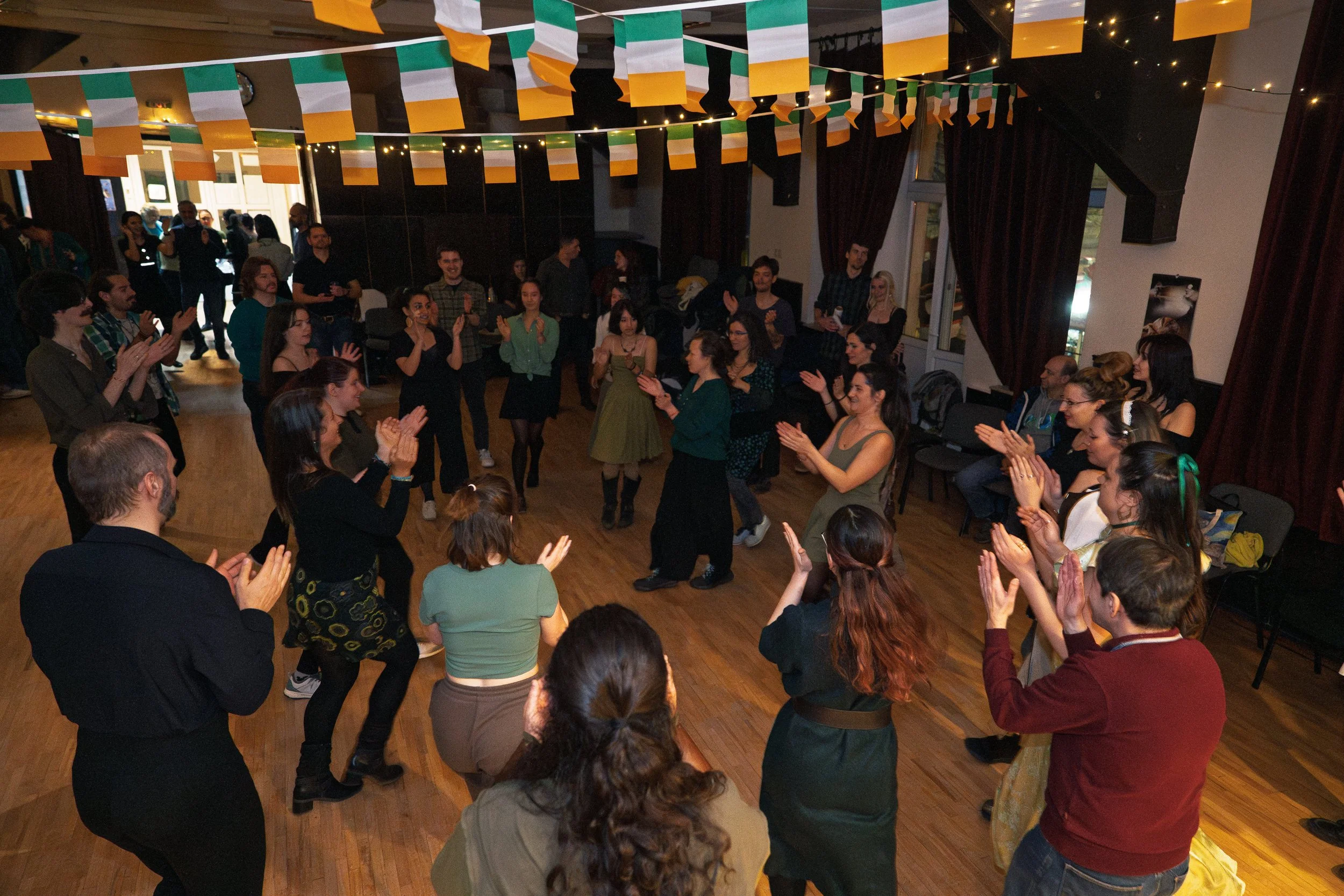 A group of people dancing and clapping in a room decorated with Irish flags and string lights, celebrating an Irish event.