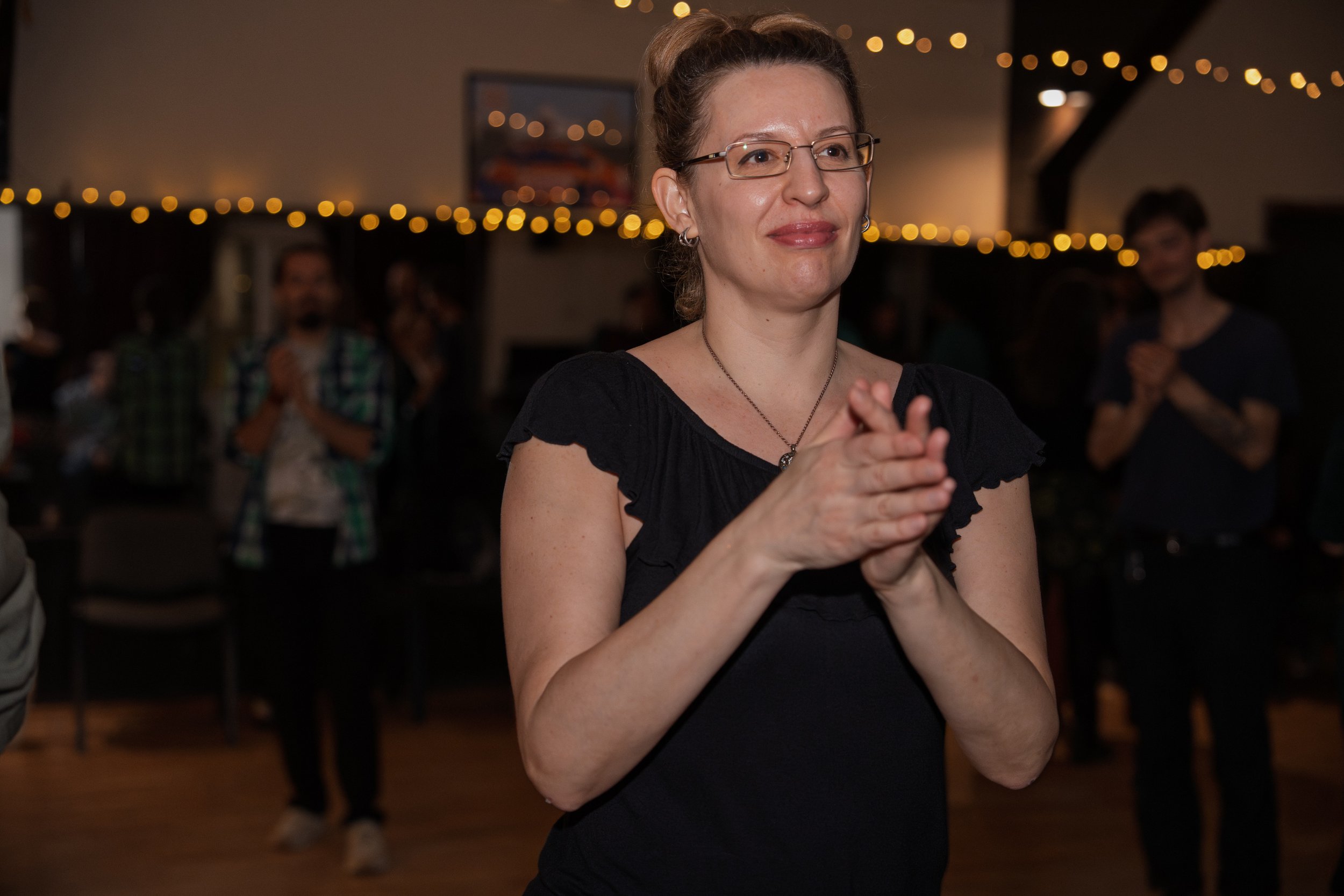 A woman with glasses and earrings claps her hands at an indoor gathering with warm lighting and blurred people in the background.