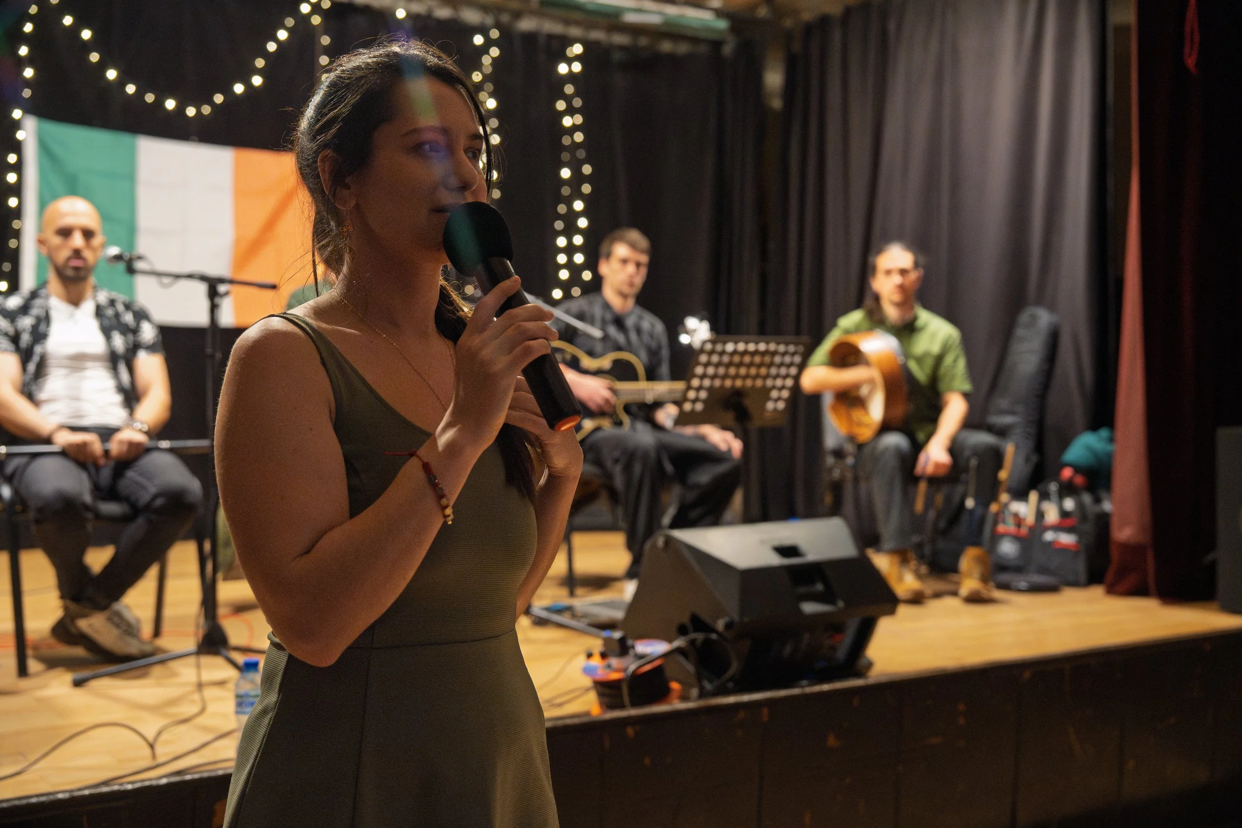 A woman holding a microphone on stage with three men in the background, two of whom are playing guitars, at a performance or event with Ireland's flag displayed behind them.