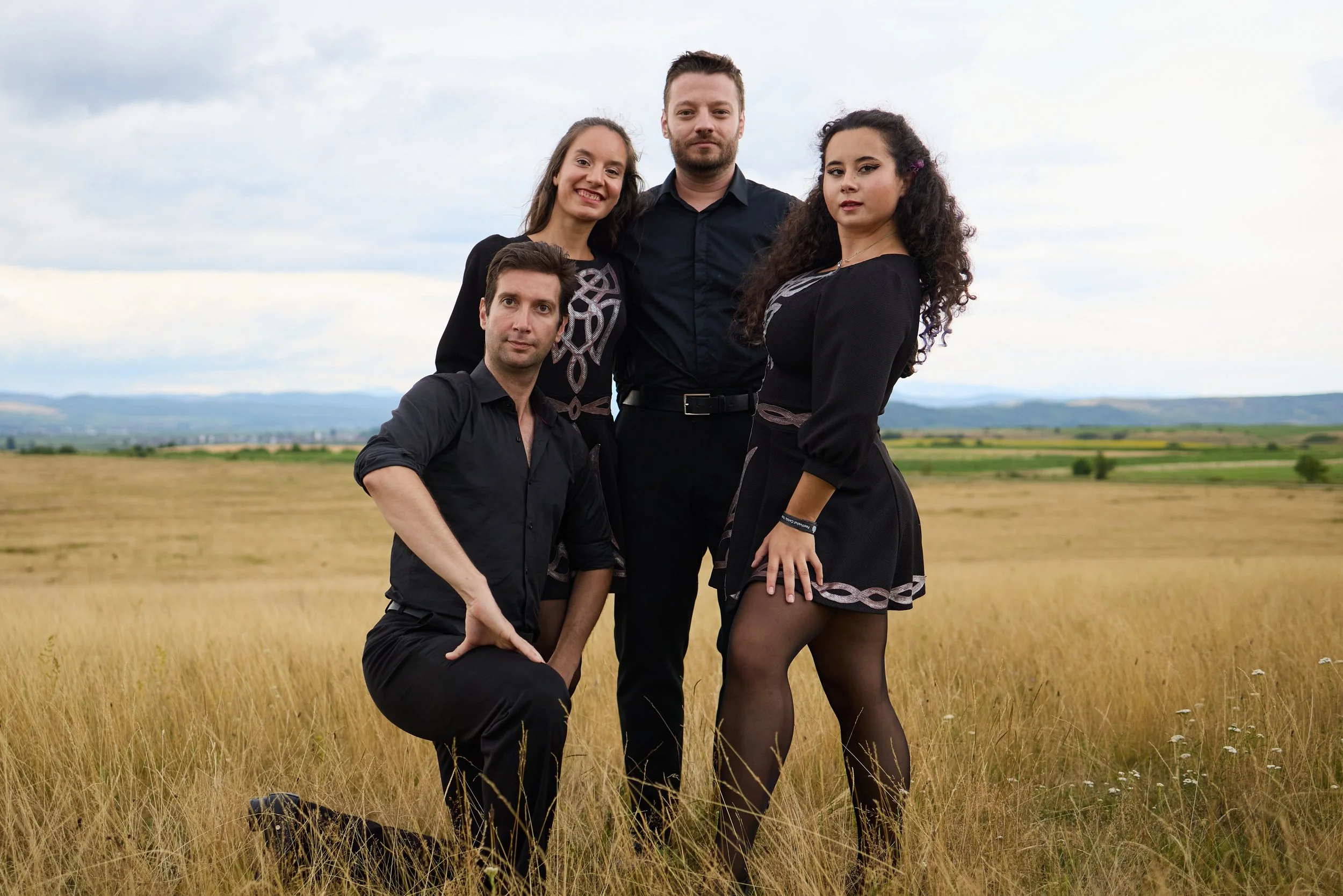 A group of five young adults posing together outdoors in a field of tall yellow grass with cloudy sky in the background.