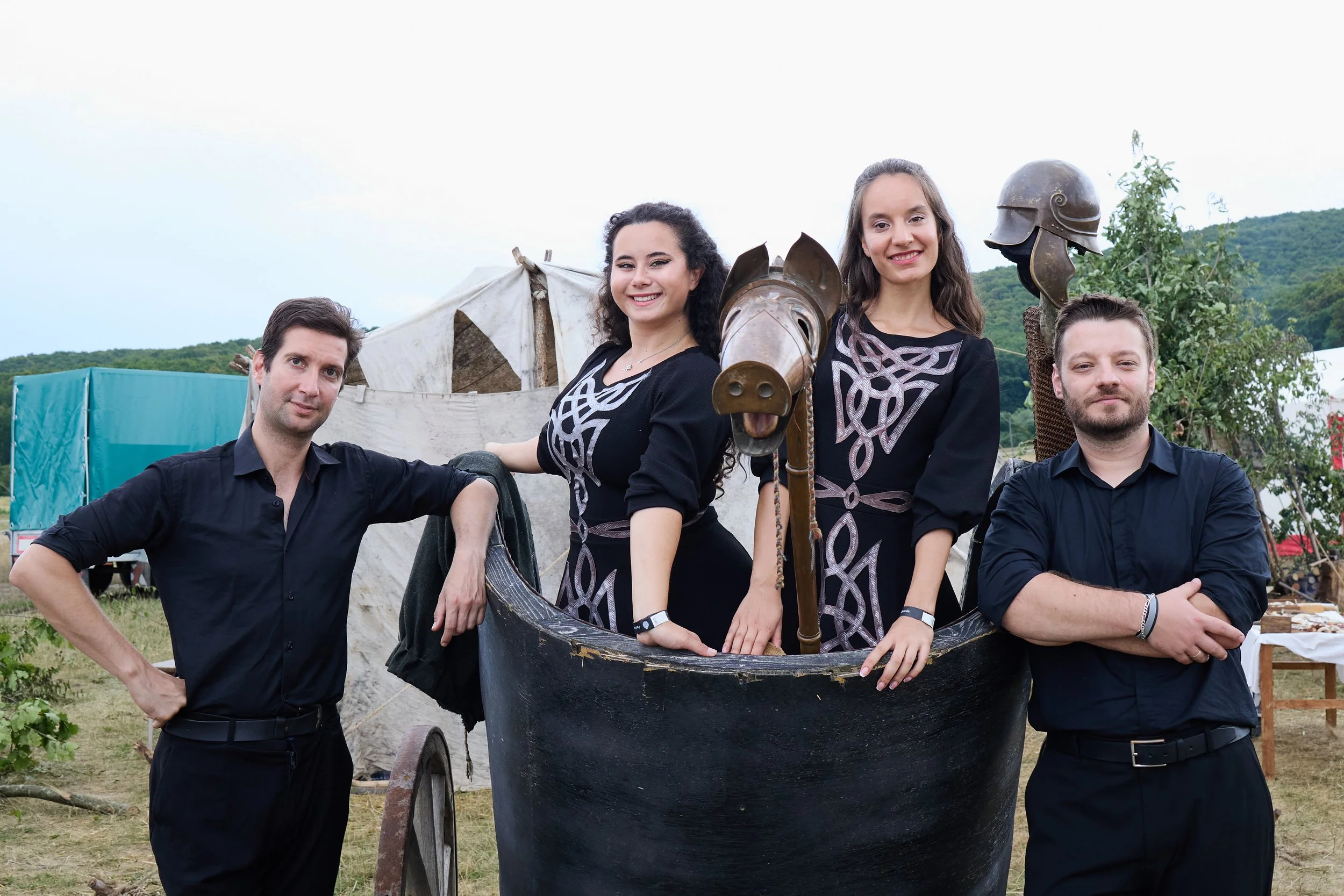 Group of four people standing outdoors in front of a historic Viking ship replica, with tents and hills in the background. Two men and two women, all smiling.