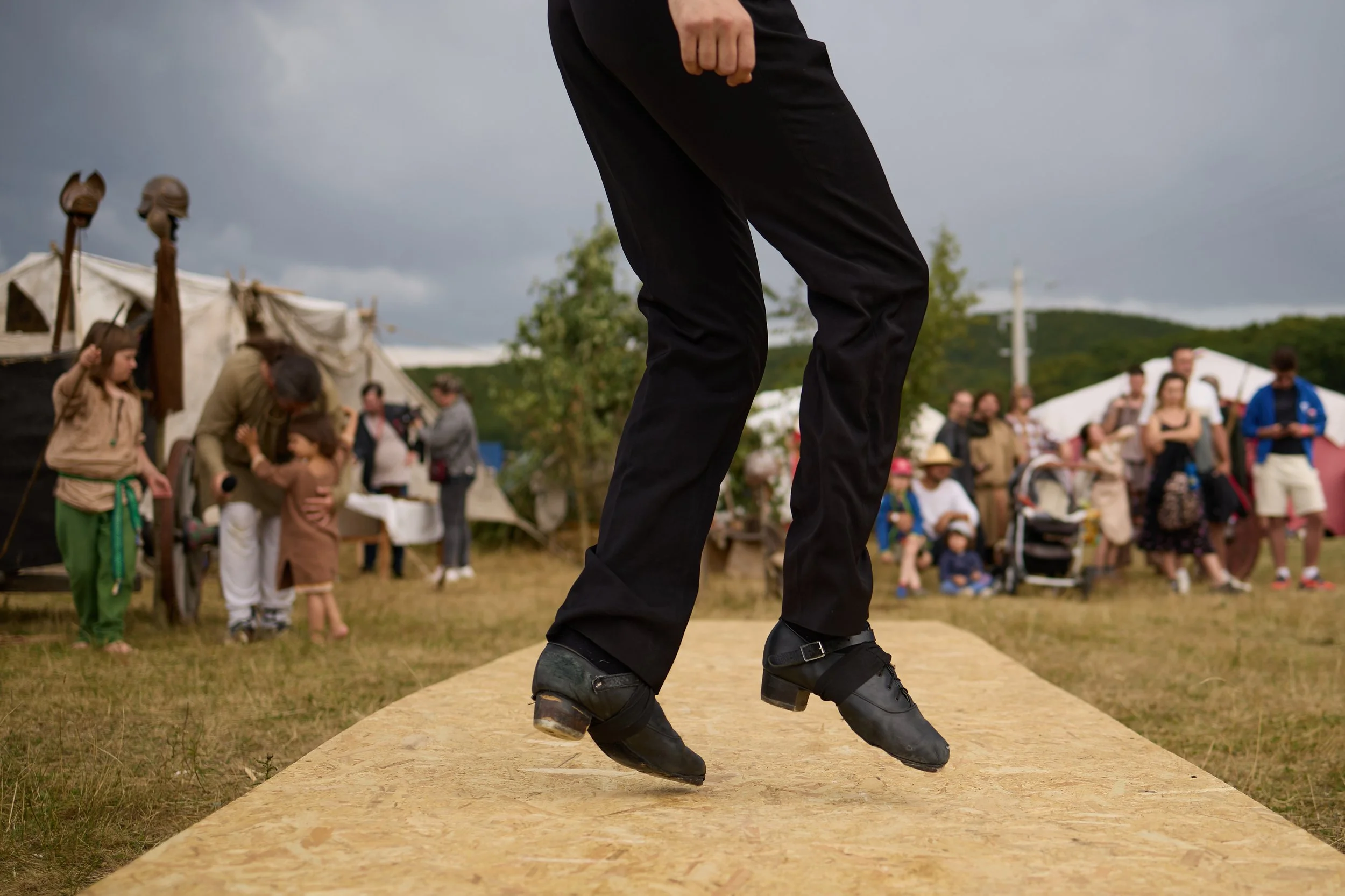 Close-up of a person standing on a wooden platform wearing black boots and black pants at an outdoor event with a crowd and tents in the background.
