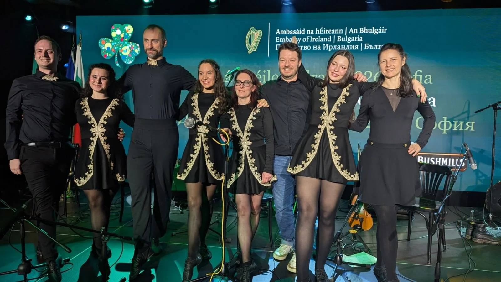 Group of performers on stage at an Irish cultural event, wearing black outfits with gold embroidery, with a backdrop displaying Irish and Bulgarian flags and the Ireland embassy logo.