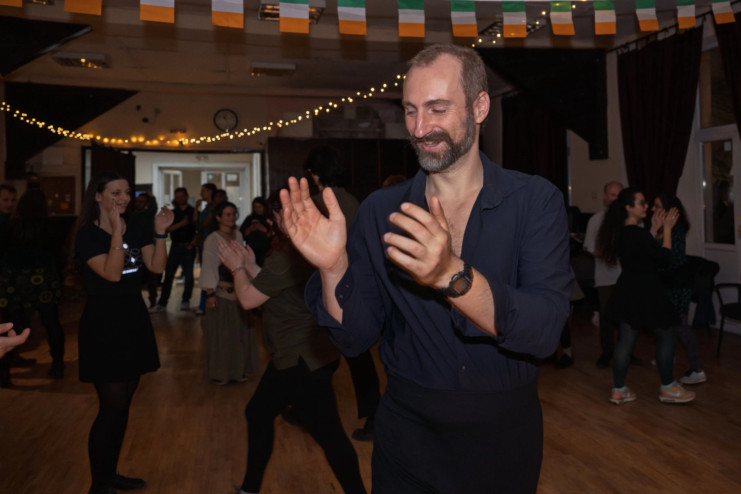 People dancing at a party or celebration in a decorated indoor venue with string lights and a clock on the wall.