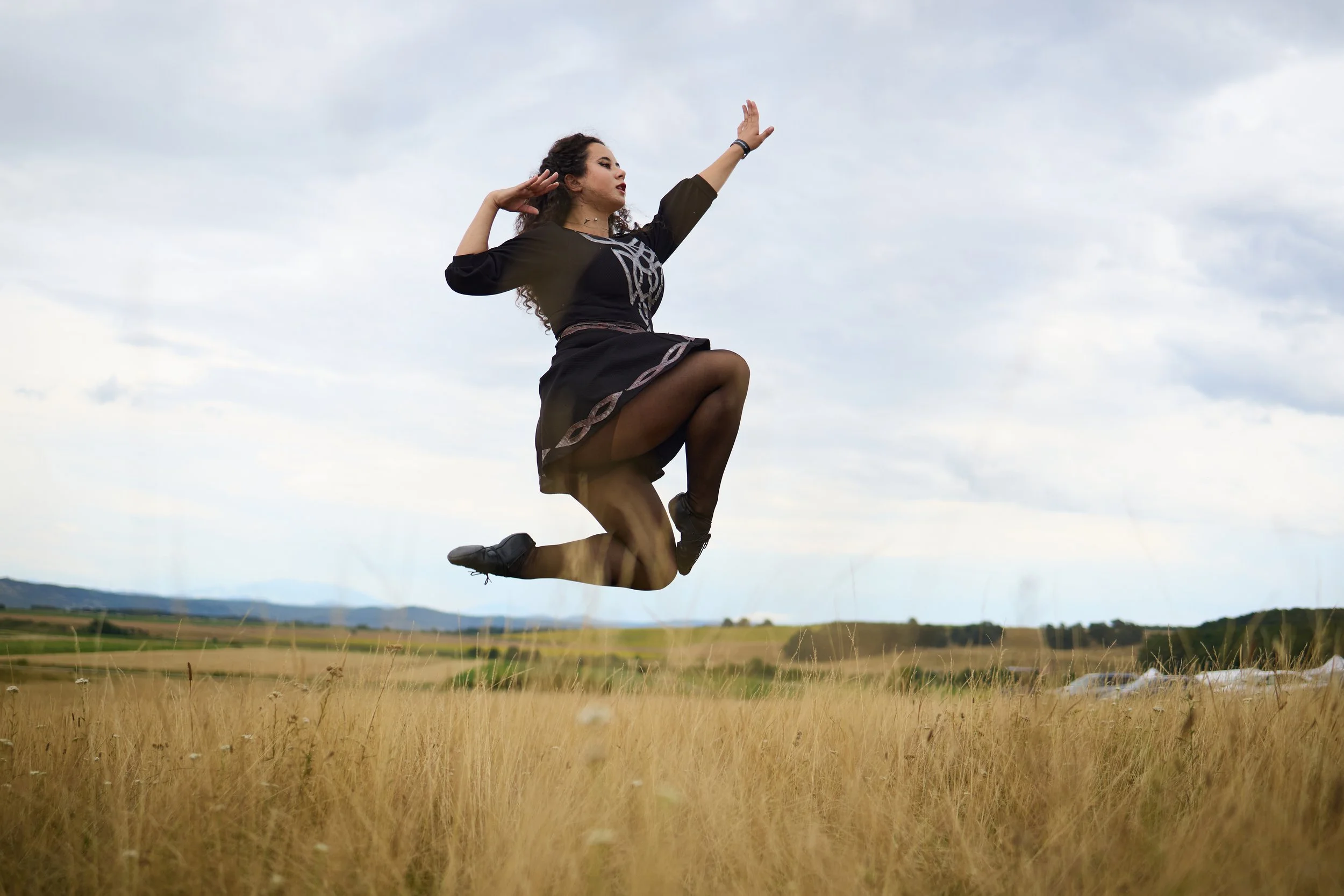 A woman in black clothing and black shoes is leaping in a grassy field with her arms raised, against a backdrop of cloudy sky and distant hills.