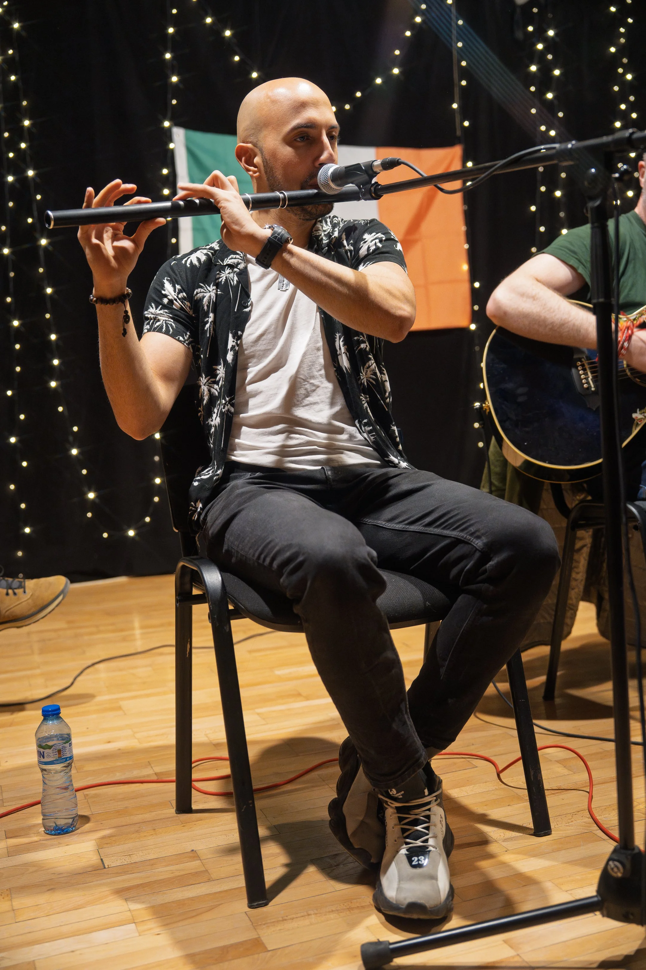 A man playing a flute while sitting on a chair, with a microphone in front of him. He is wearing a black Hawaiian shirt with white palm tree patterns, black pants, and sneakers. There is a water bottle on the wooden floor near his feet, and another p