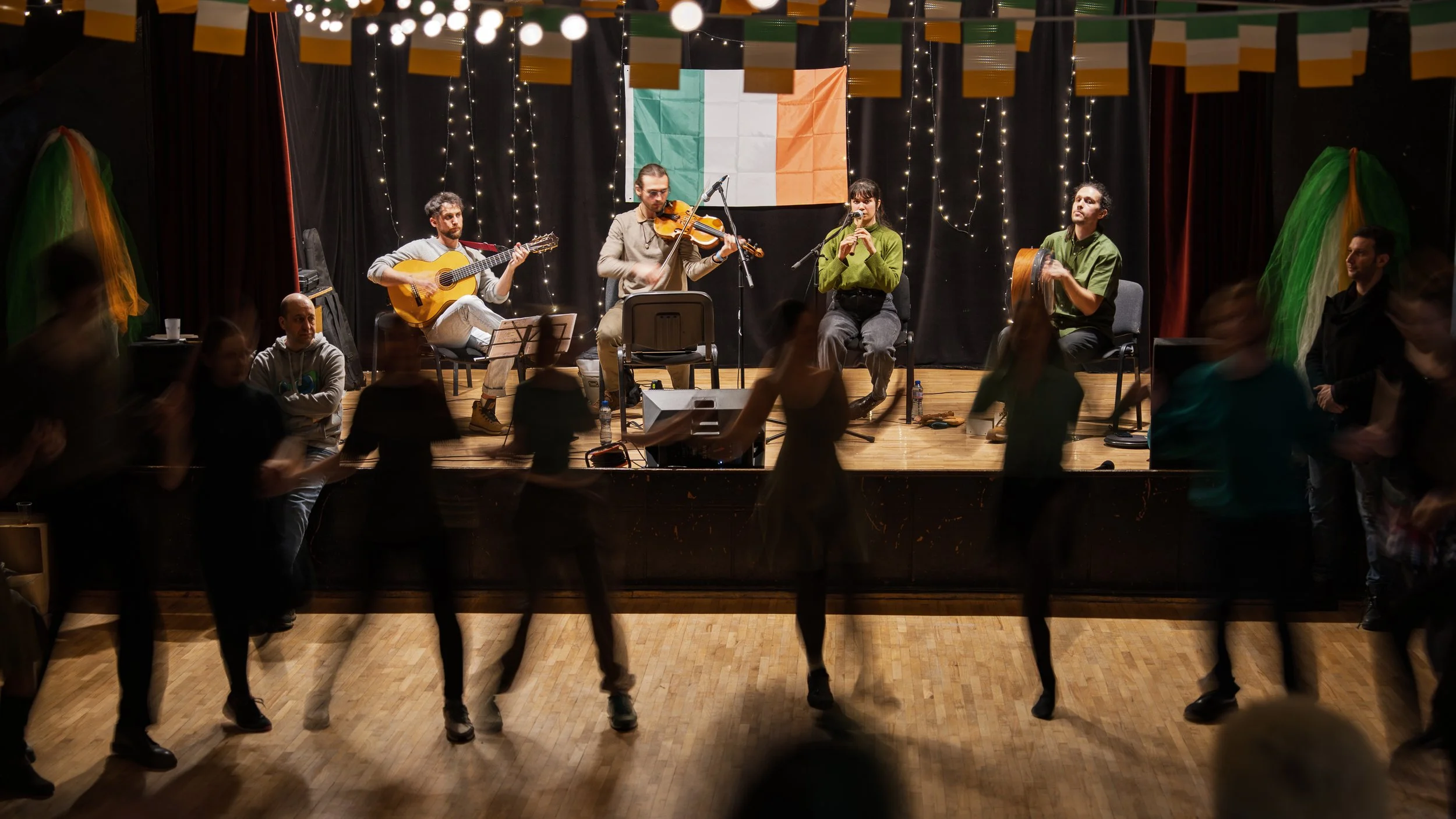 A live Irish music performance on a stage with musicians playing guitar, violin, flute, and percussion. The Irish flag hangs in the background, and people are dancing in front of the stage.