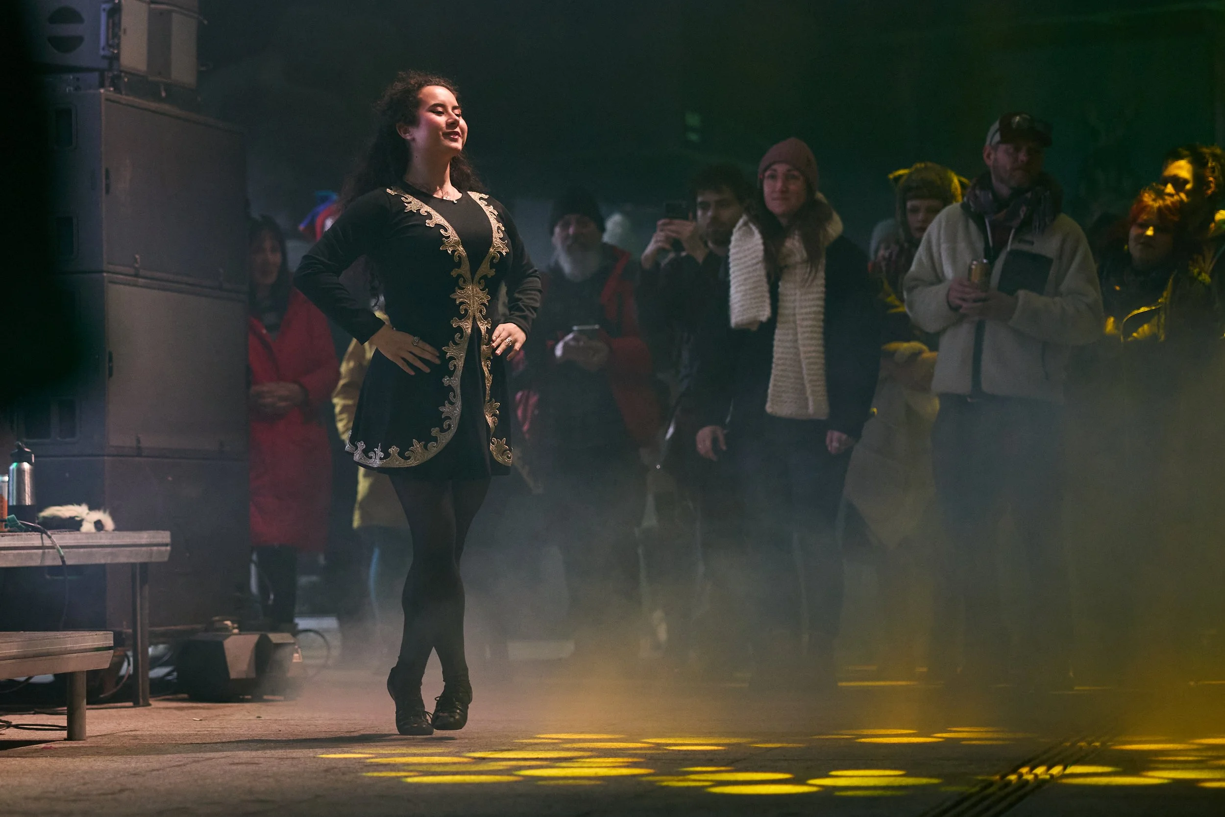 A woman standing on a stage with her hands on her hips, wearing a black dress with gold embroidery, in front of a diverse audience in a dimly lit venue.