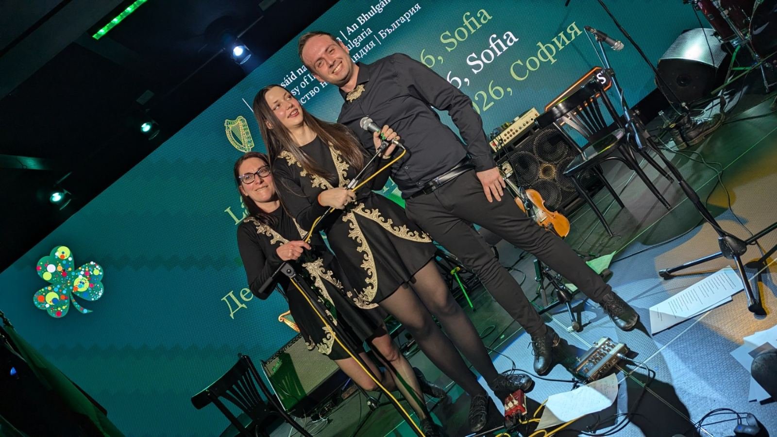Three performers standing on stage with musical instruments, smiling and posing for a photo, behind large screen with text in multiple languages and a colorful logo.