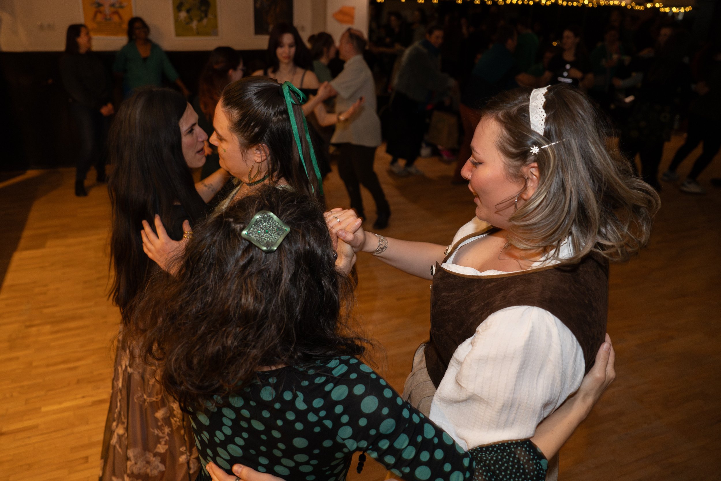 Two women are dancing closely on a wooden floor, holding hands and smiling at each other. Other couples dance in the background at a lively indoor event, with artwork on the walls.