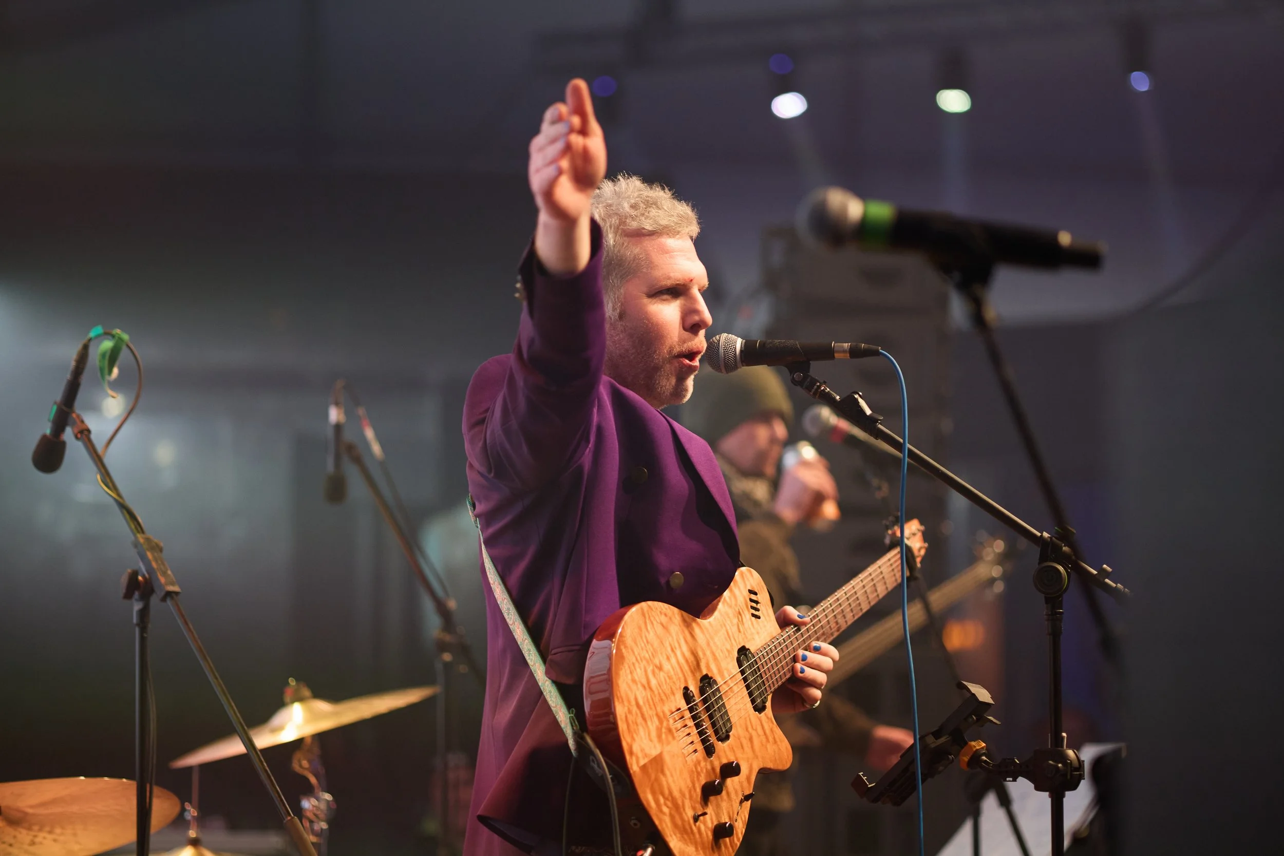 A man in a purple jacket playing an acoustic guitar and singing into a microphone on stage during a live performance.