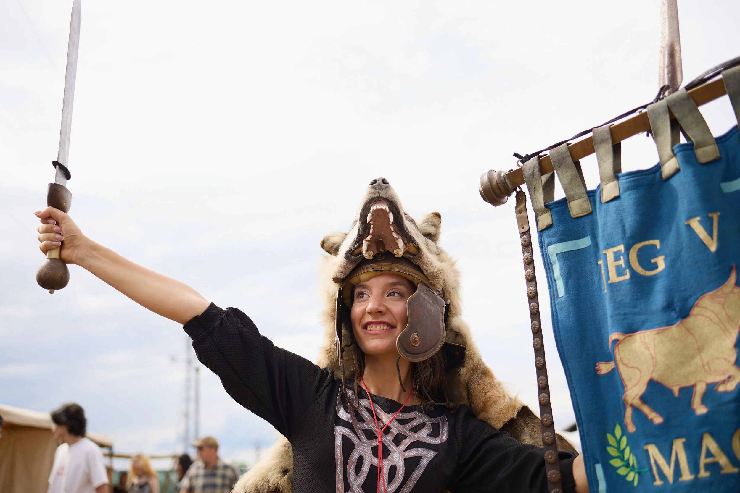 A woman wearing a furry hat with a wolf head and open mouth, smiling as she raises her arm, holding a staff, during an outdoor event with many people in the background.