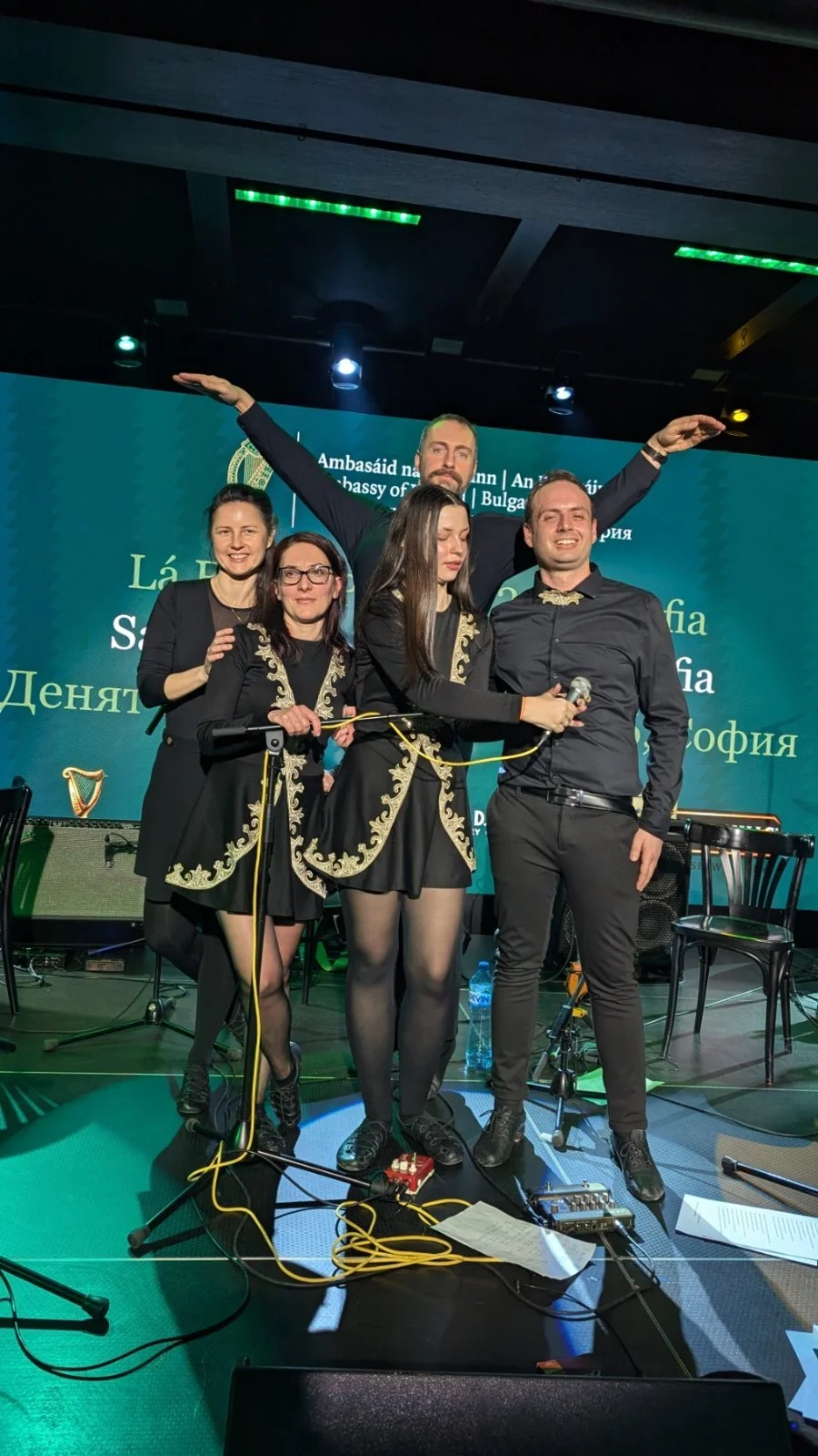 Five performers on stage at a formal event, three women and two men. The women are holding a microphone, dressed in black outfits with gold embroidery, and the men are in black shirts and pants. They are smiling and posing for the photo with musical 