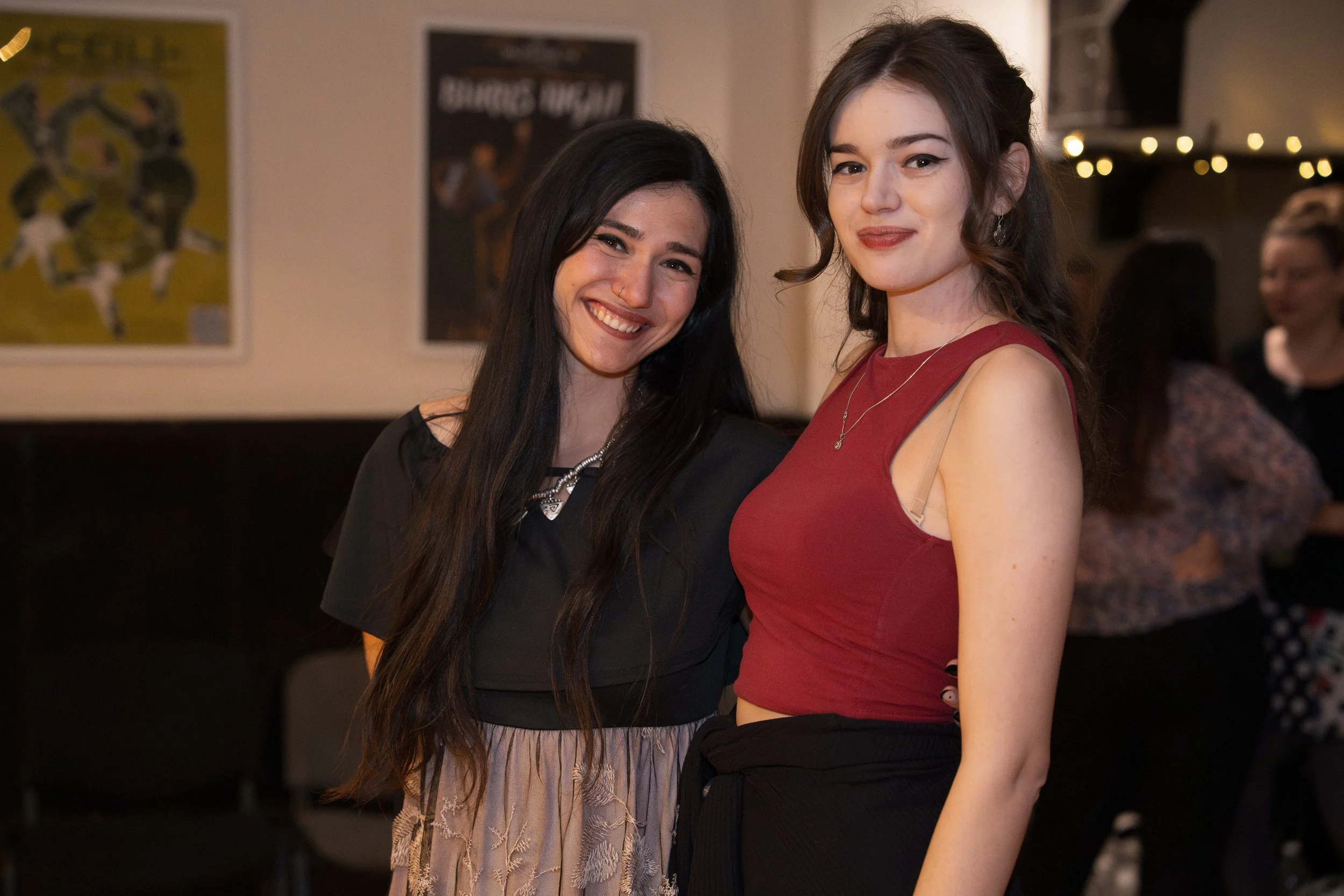 Two young women smiling and posing together at an indoor event, with artwork on the wall behind them and string lights in the background.