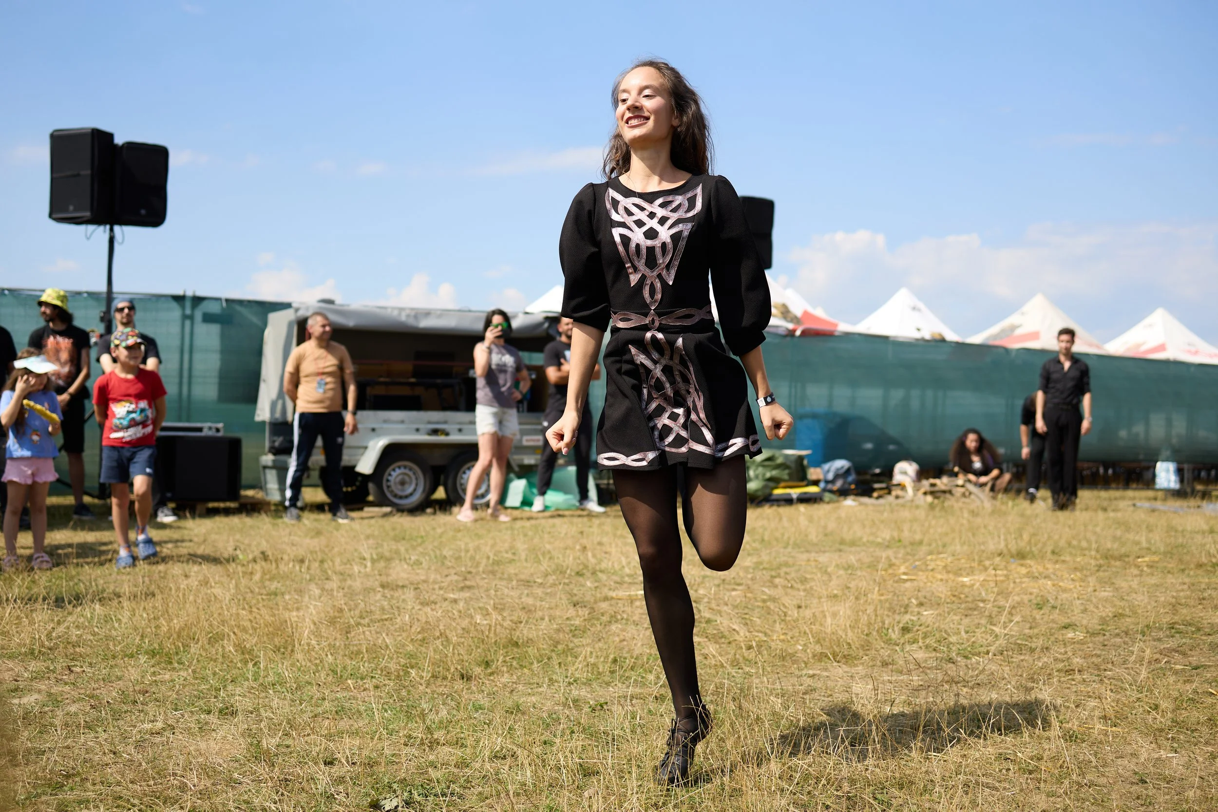 A woman in black tights and a black dress with pink and black design runs across a grassy field at an outdoor event. There are onlookers in the background, tents, and blue sky.