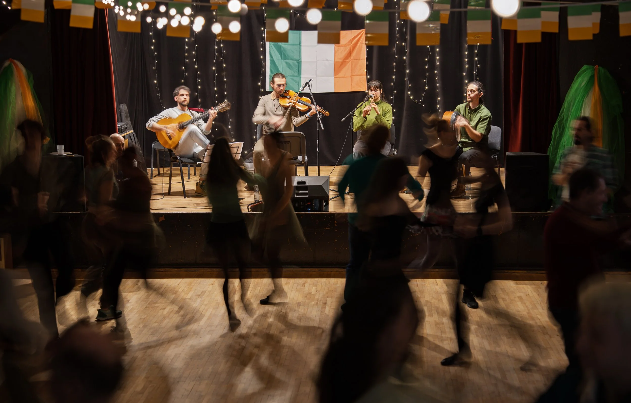 A musical performance on a stage with five musicians, including guitarists and a flutist, during a lively dance event. The backdrop features the Irish flag.