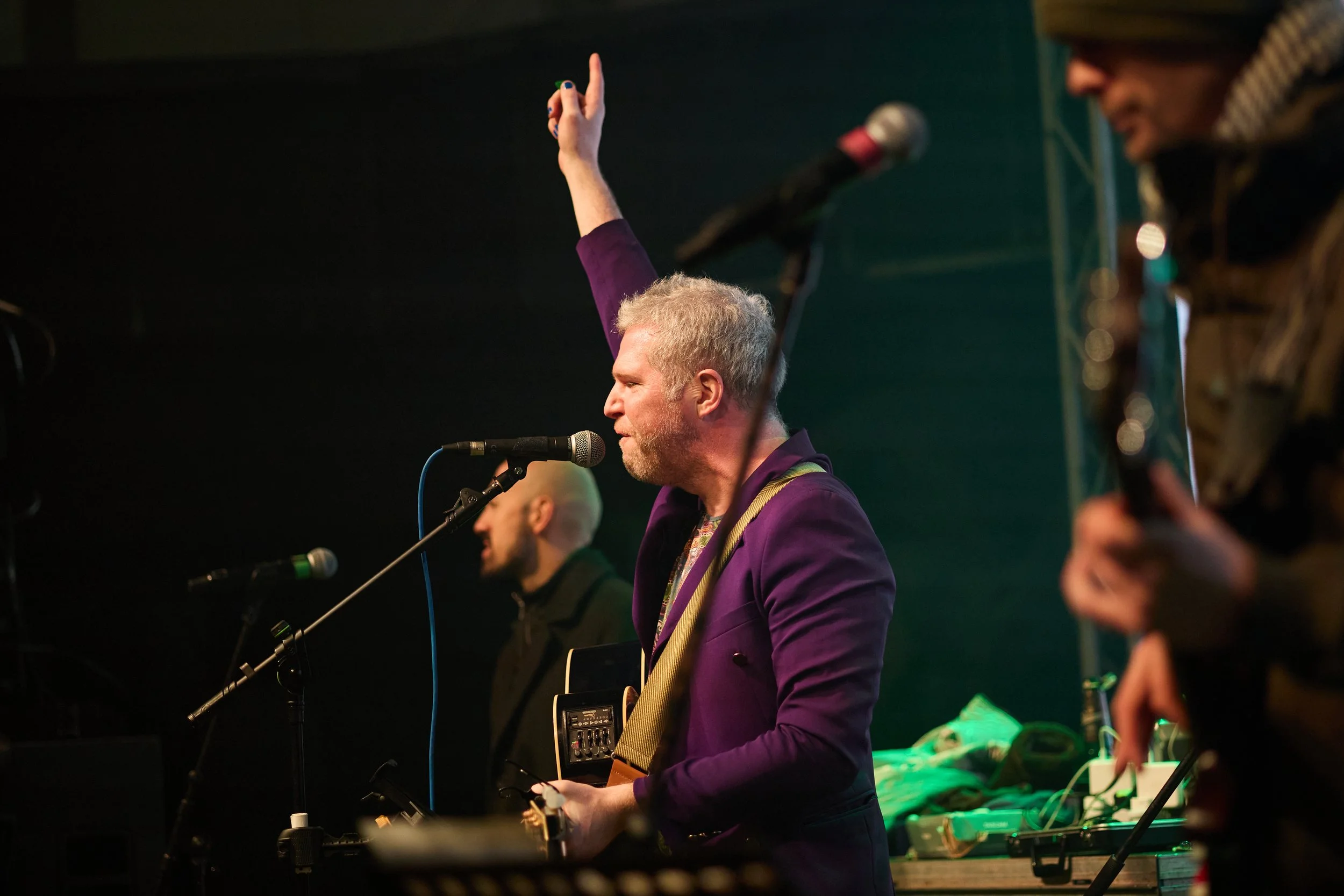 A man with gray hair and a beard, singing into a microphone while playing a guitar at a live music performance, with other band members in the background.