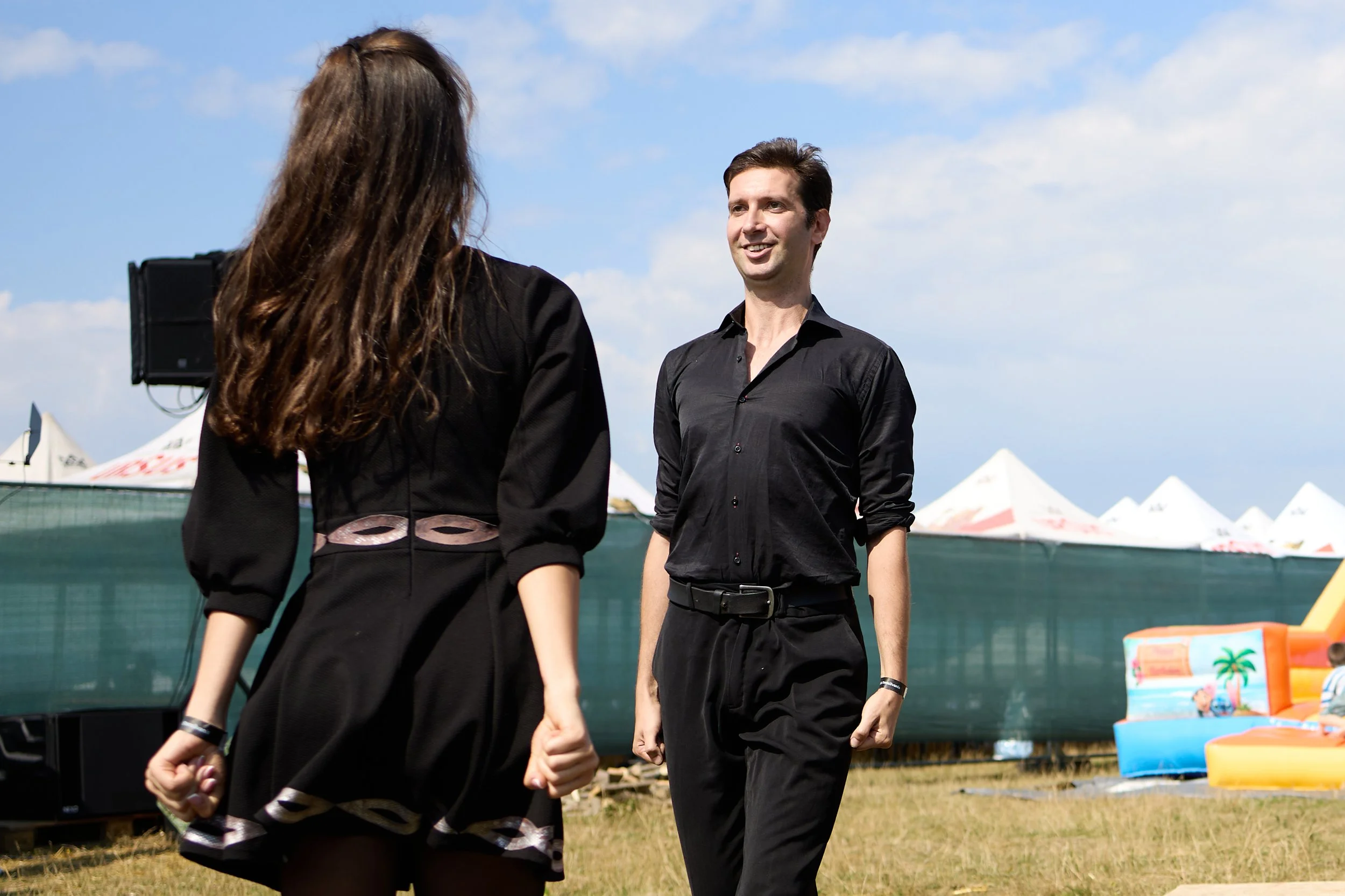 A man and a woman are outside at a daytime event, with the woman facing away and the man smiling at her. The woman has long brown hair and is wearing a black dress, while the man has short dark hair and is dressed in black. There are tents and colorf